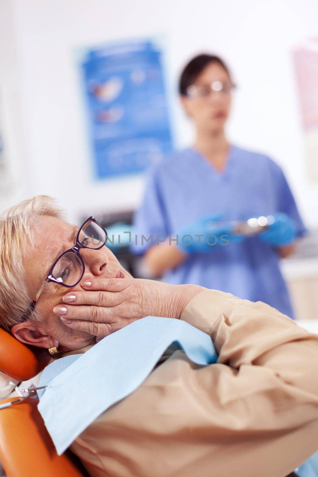 Middle age patient touching mouth with painful expression sitting on chair in dentist cabinet. Senior woman in healthcare hospital accusing and complaining about tooth.