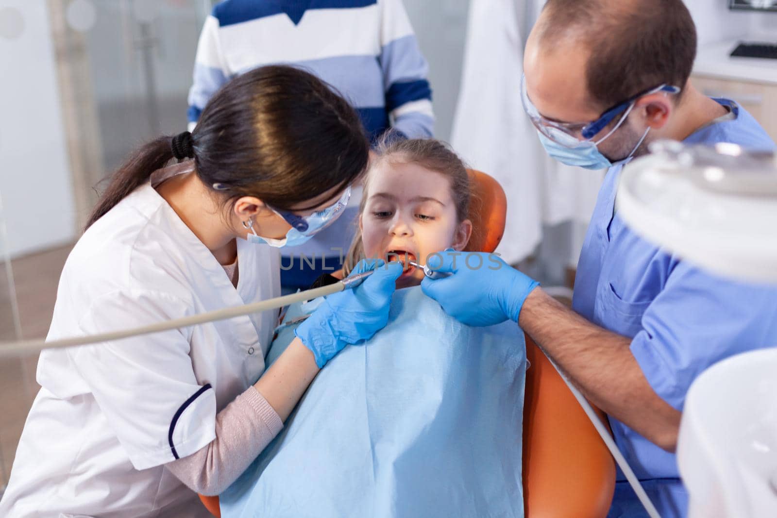 Little girl with mouth open in the course of cavity treatment sitting on dental chair. Mother with her kid in stomatology clinic for teeth examine using modern instruments.