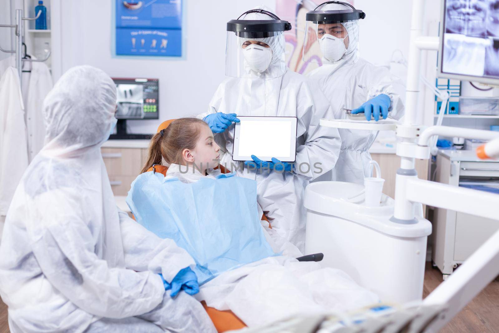 Dentist doctor in ppe suit talking about dental cavity to little girl and her mothern. Medical specialist in oral hygine holding tablet pc with copy space available during global pandemic with covid19.