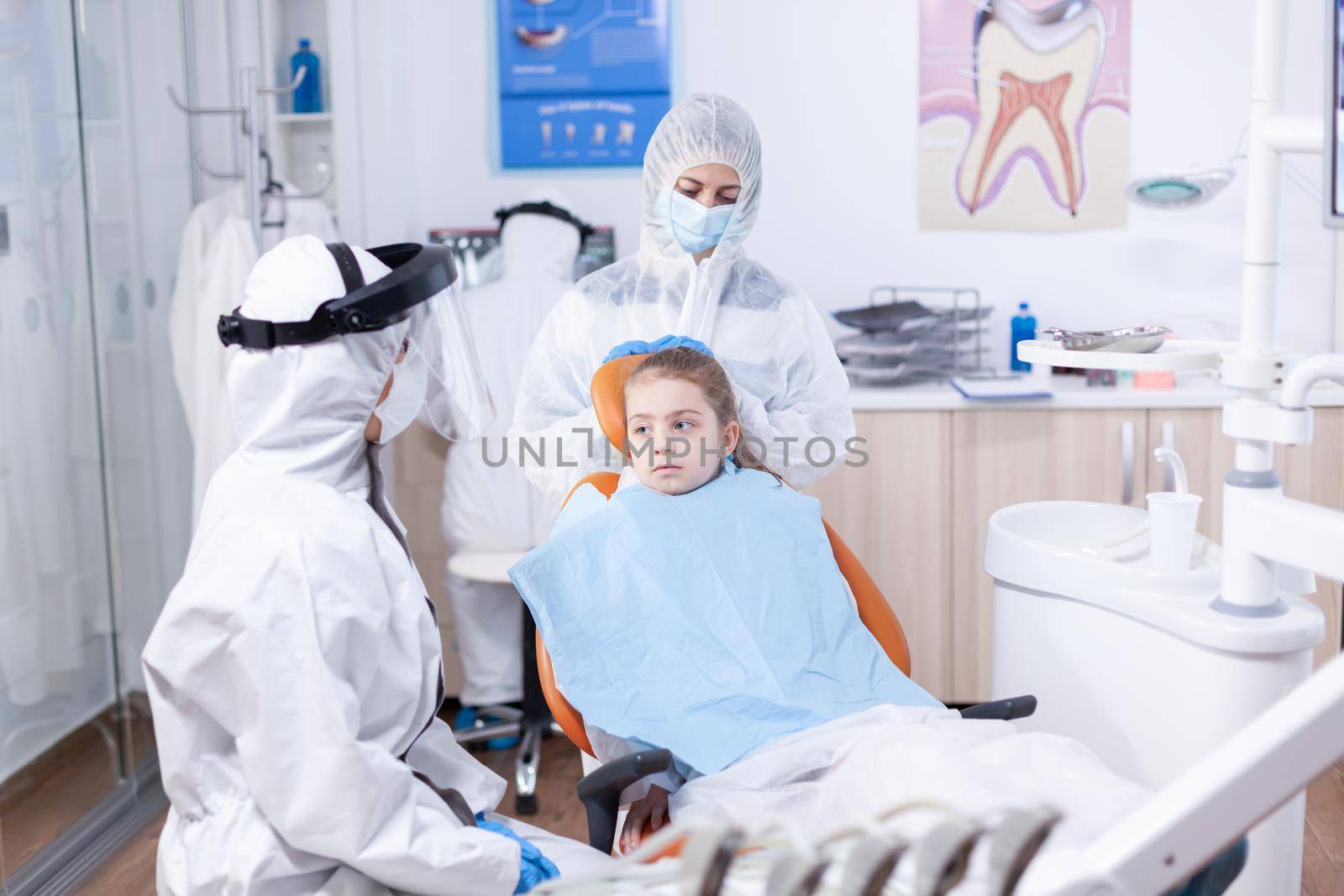 Dentist in coverall talking with kid before dentistiry examination with face mask. Stomatologist during covid19 wearing ppe suit doing teeth procedure of child sitting on chair.