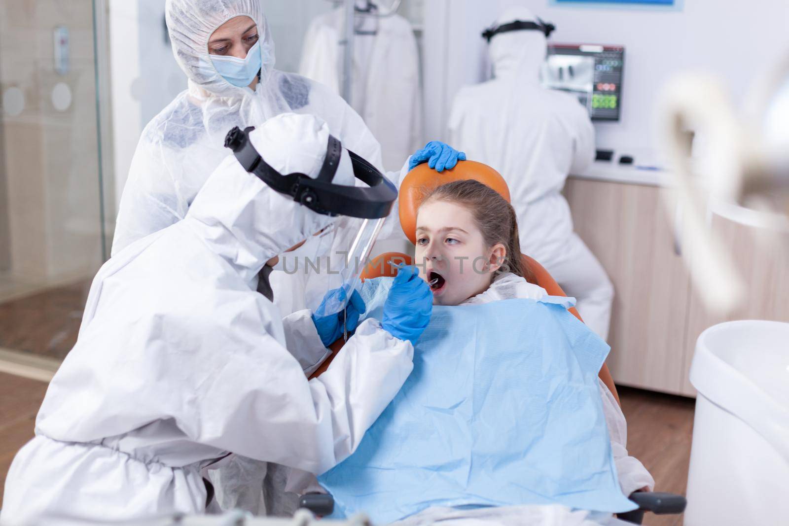 Stomatolog in ppe suit during teeth examination of child wearing bib sitting on dental chair. Dentist in coronavirus suit using curved mirror during teeth examination of child.