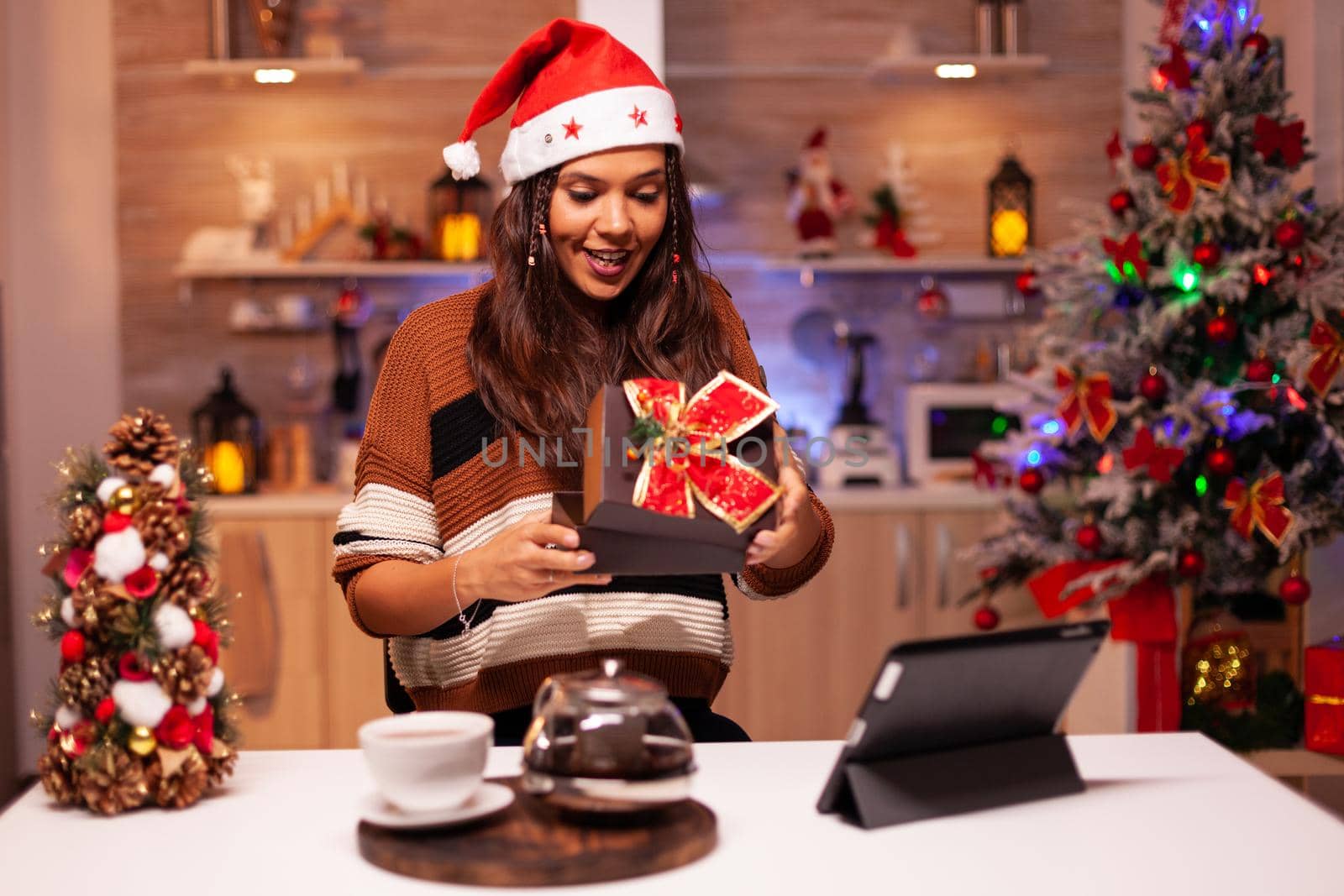 Happy woman opening gift box while on online video call conference with family using tablet. Caucasian adult with santa hat receiving present feeling cheerful at christmas decorated home