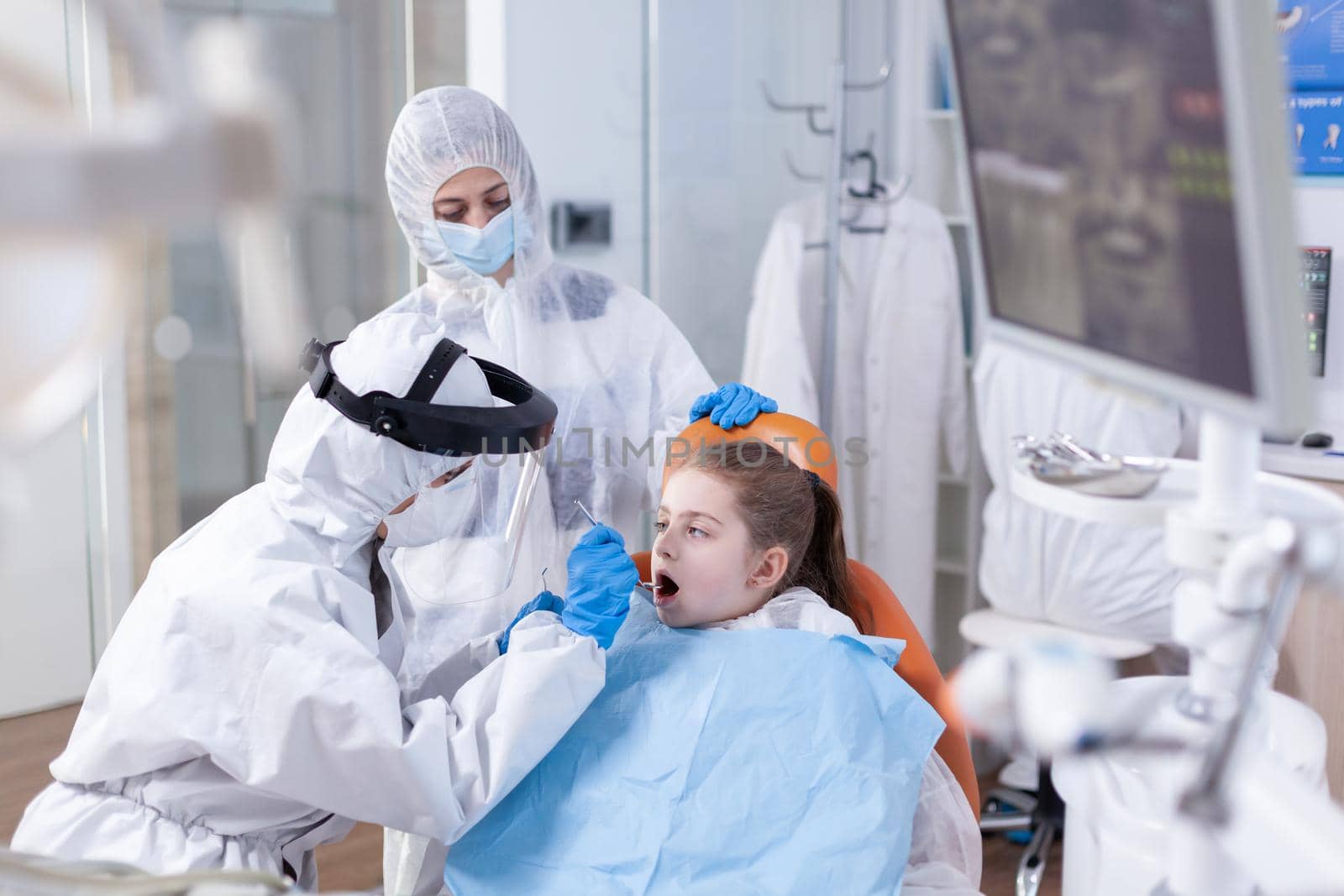 Stomatolog in the course of caries treatment for kid dressed in ppe suit as safety precaution Dentist in coronavirus suit using curved mirror during teeth examination of child.