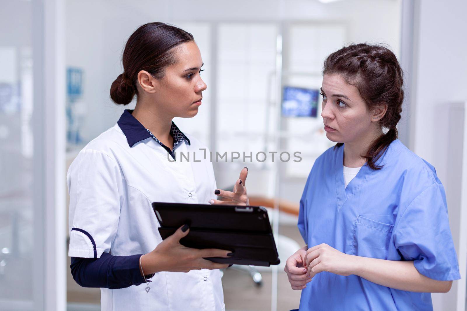 Dentist looking at nurse explaining teeth infection holding tablet pc. Orthodontic specialist and assisant at reception in dental clinic discussing about teeth treatment.