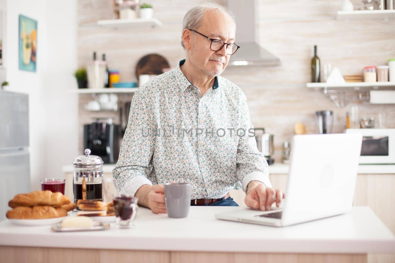 Senior man smiling and using laptop in the kitchen. Daily life of senior man in kitchen during breakfast using laptop holding a cup of coffee. Elderly retired person working from home, telecommuting using remote internet job online communication on modern technology notebook