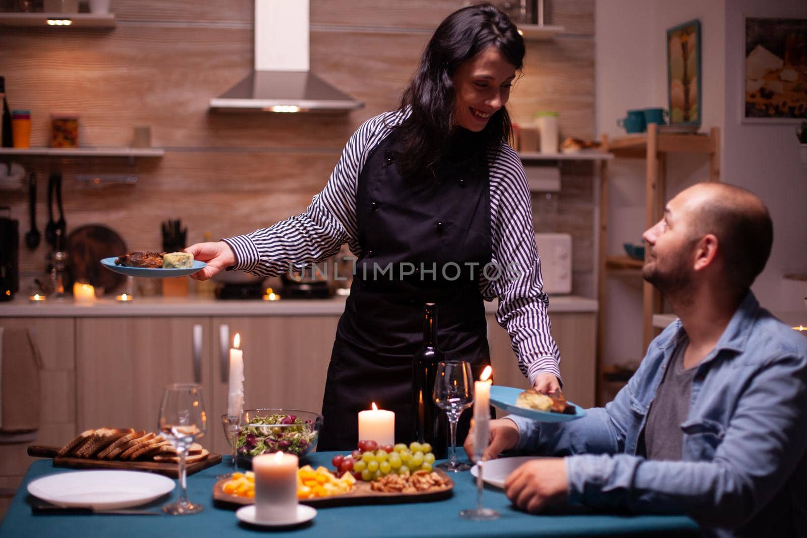 Wife surprising husband with dinner in dining room and celebrating with delicious food. Woman preparing festive dinner, cooking for his man romantic dinner, talking, sitting at table