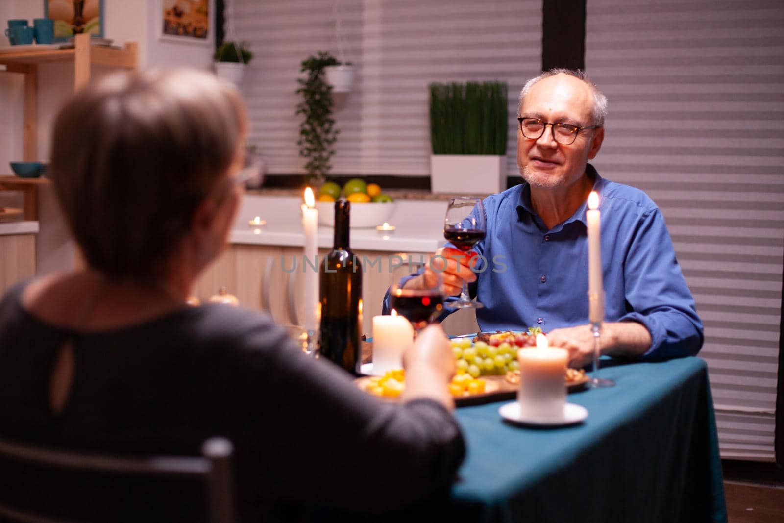 Senior man telling a story to his wife while celebrating in kitchen with wine and food. Senior couple sitting at the table in dining room , talking, enjoying the meal, celebrating their anniversary in the dining room.