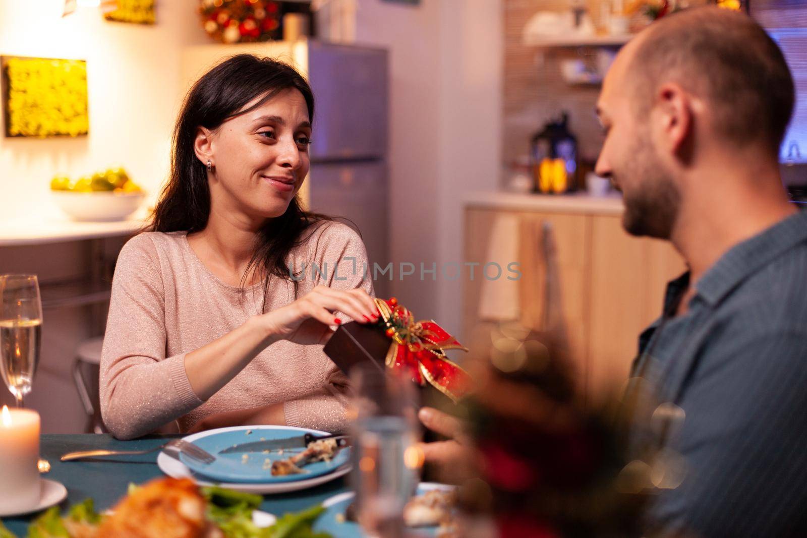 Happy couple enjoying christmas holiday surprising with xmas present with ribbon on it sitting at table in x-mas decorated kitchen. Cheerful family celebrating winter holiday spending time together