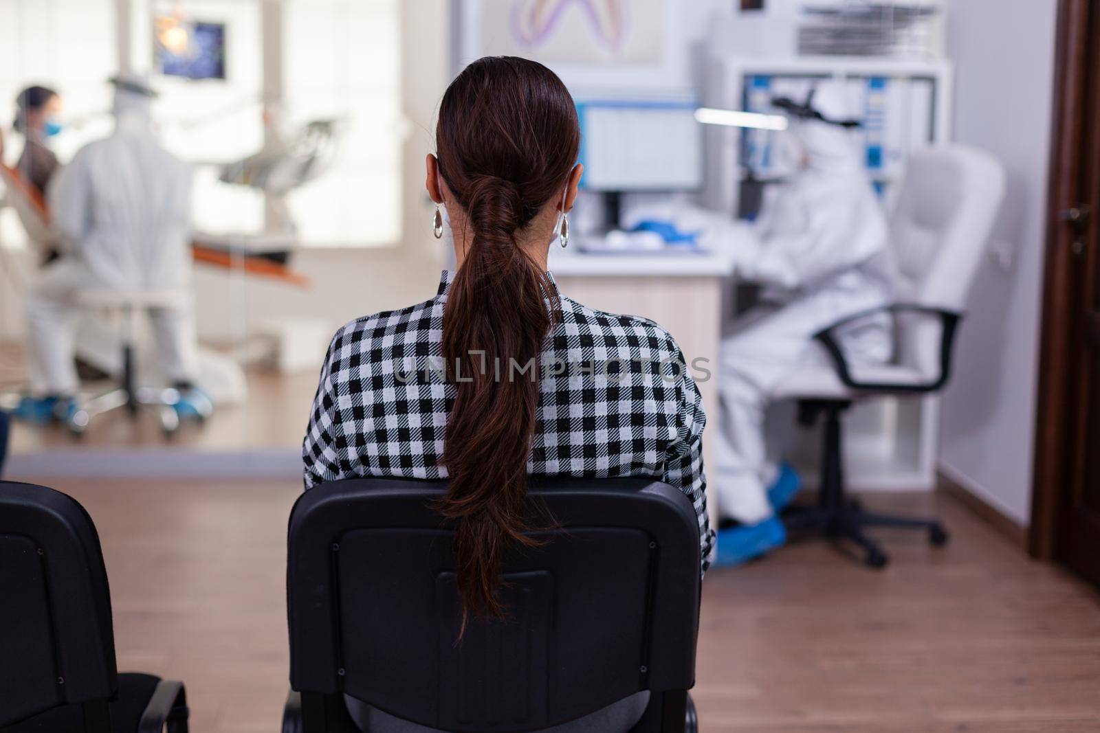 Back view of woman in dentist office waiting for consultation sitting in chairs on waiting room, assitant in ppe suit filling paper work on computer, dentistiry doctor consulting patient.