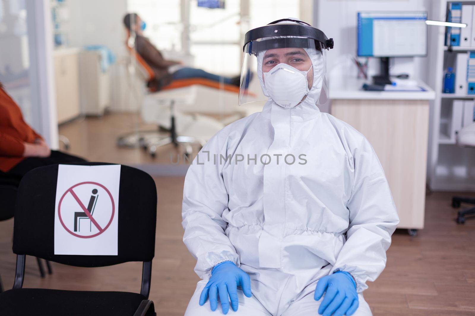Close up of tired doctor in stomatological office looking on camera wearing overall and face shield sitting on chair in waiting room clinic. Concept of new normal dentist visit in coronavirus outbreak.