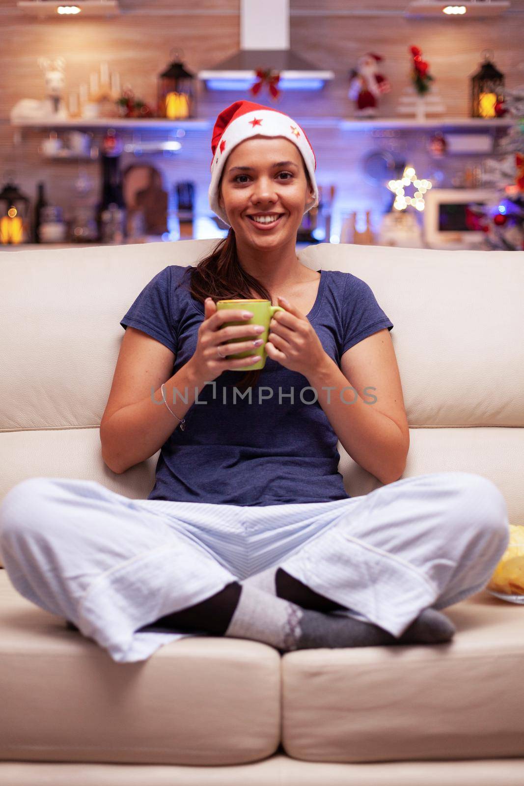 Portrait of smiling woman wearing red santa hat sitting in lotus position on sofa in xmas decorated kitchen looking into camera enjoying christmas holiday. Woman celebrating winter season