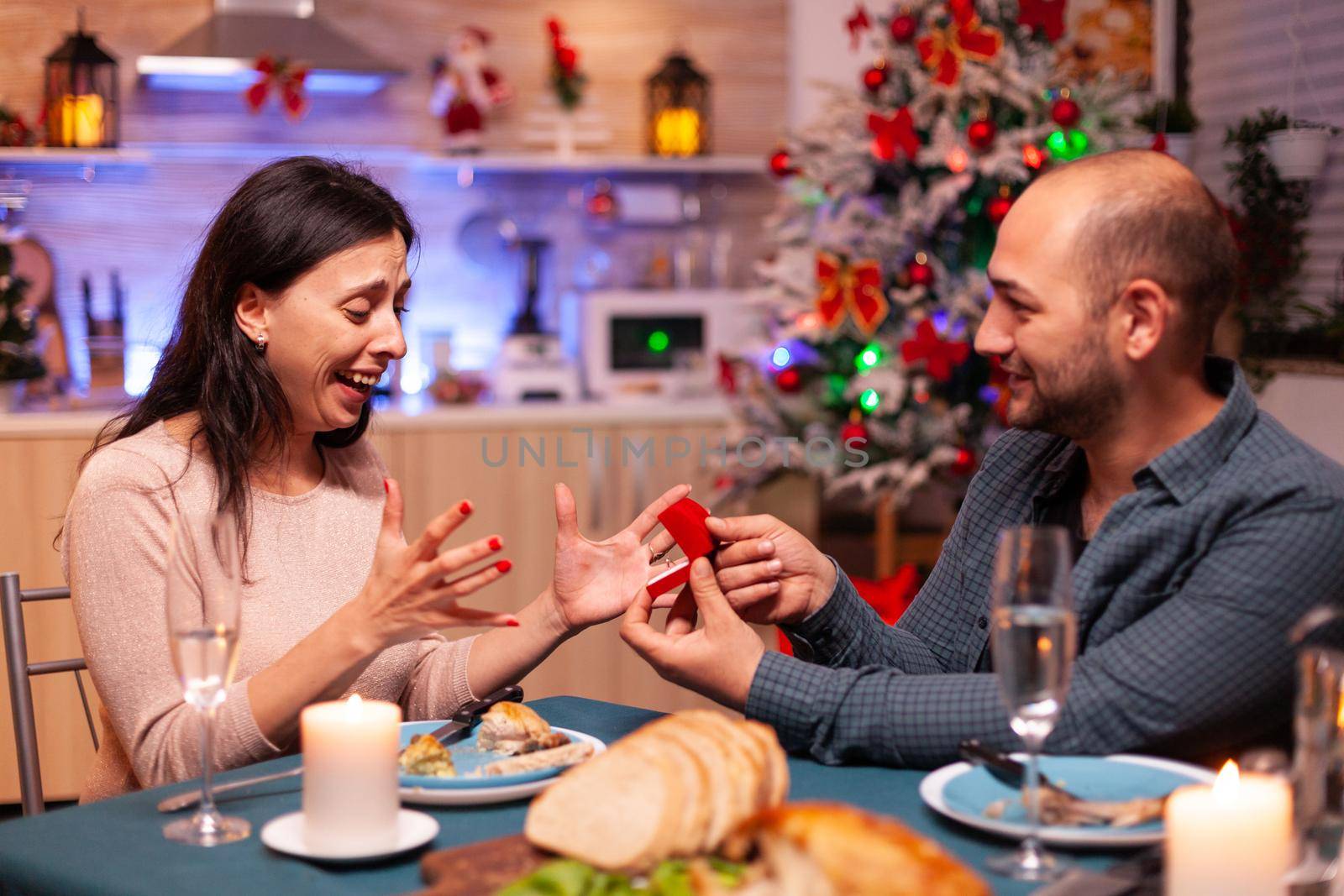 Happy man surpsing girlfriend with diamond luxury expensive engagement ring celebrating christmas holiday together sitting at dining table in xmas kitchen. Couple enjoying winter season