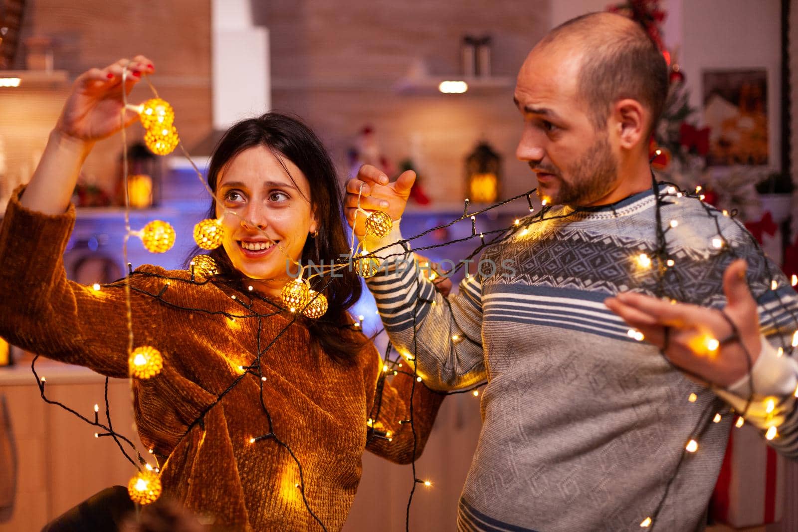 Happy amused married couple stucking in christmas tree lights enjoying spending winter holiday together. Cheerful family decorating kitchen celebrating christmastime. Santa-claus tradition