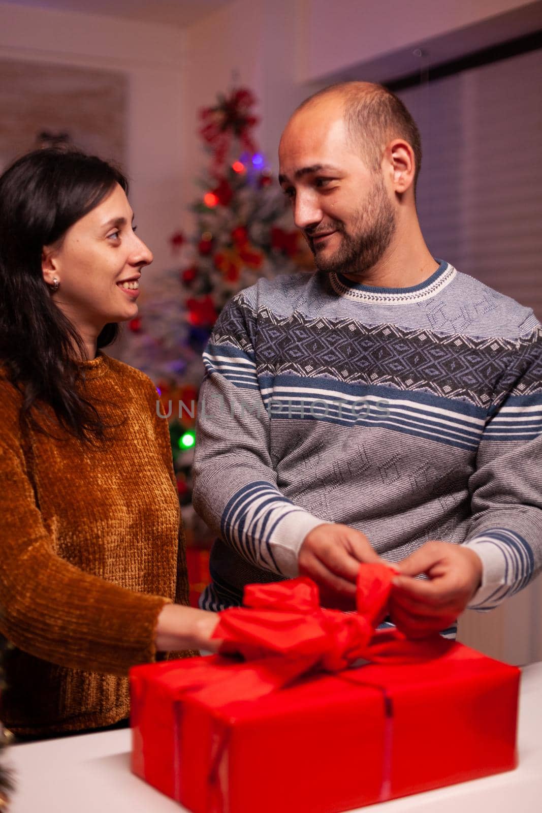 Cheerful family opening xmas decorated gift with ribbon on it during christmas holiday standing in x-mas kitchen. Happy couple married celebrating winter season. Santa-claus tradition