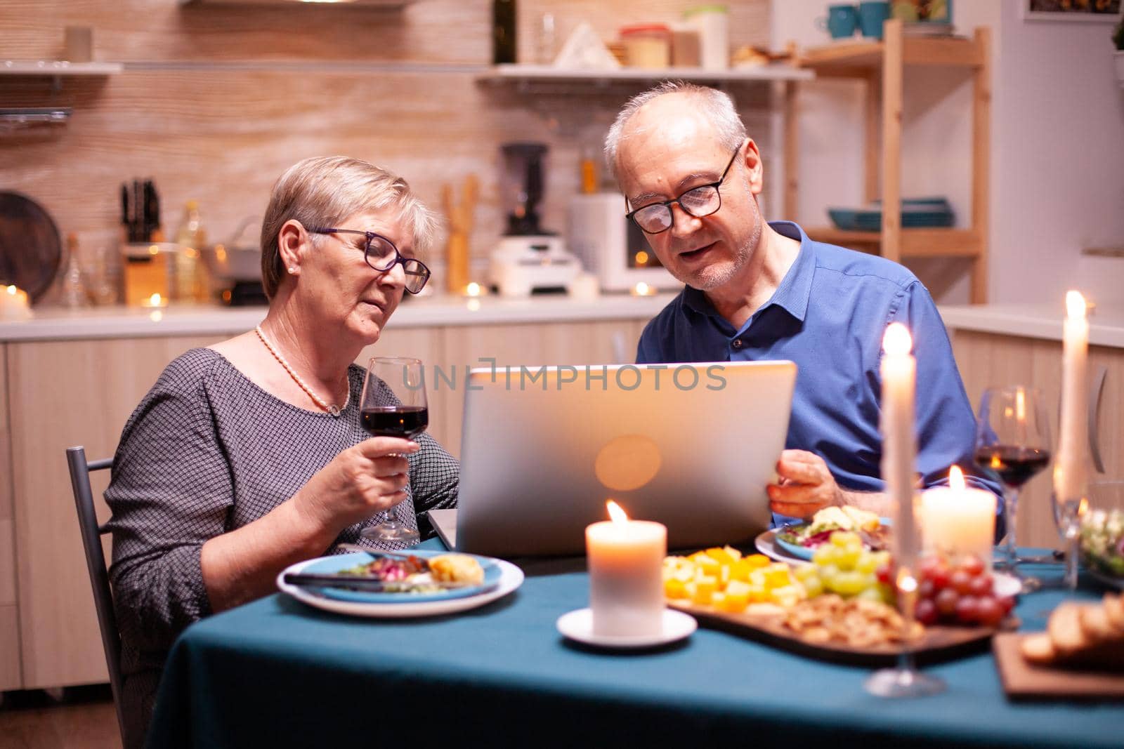 Elderly couple using laptop in the kitchen celebrating relationship. Senior people sitting at the table browsing, searching, using laptop, technology, internet, celebrating their anniversary in the dining room.