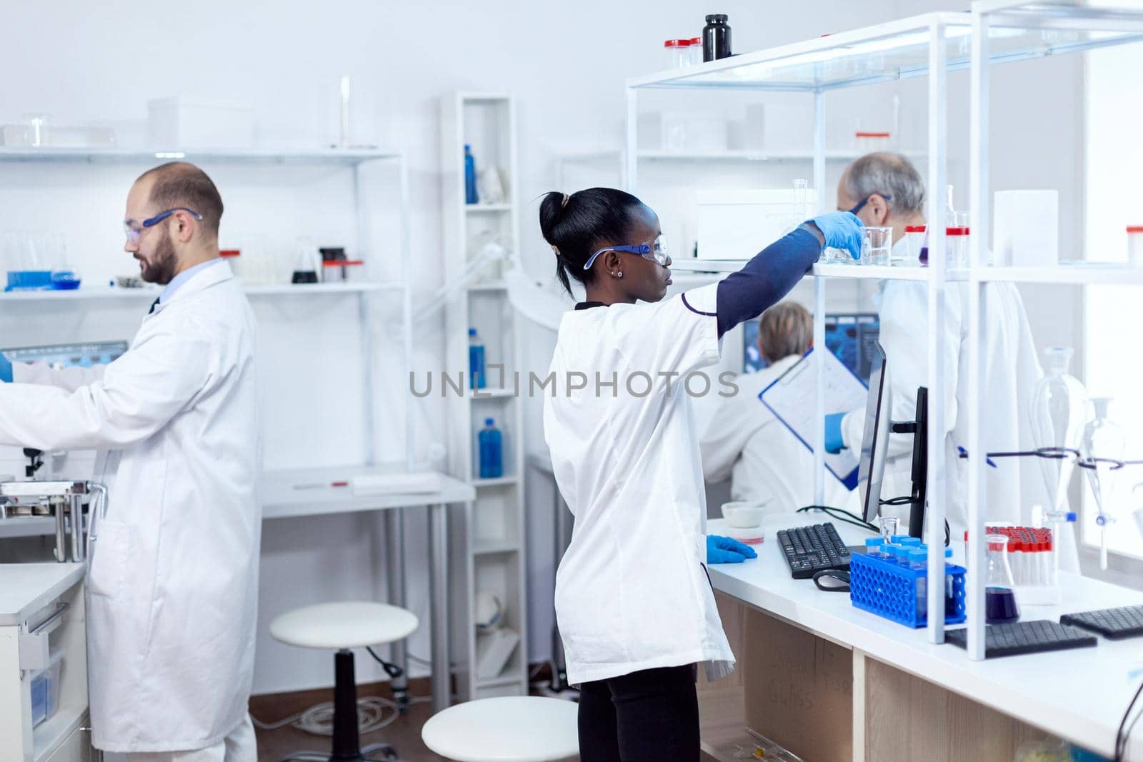 African chemist in professional laboratory taking a glass flask with solution from shelf. Multiethnic team of researchers working in microbiology lab testing solution for medical purpose.