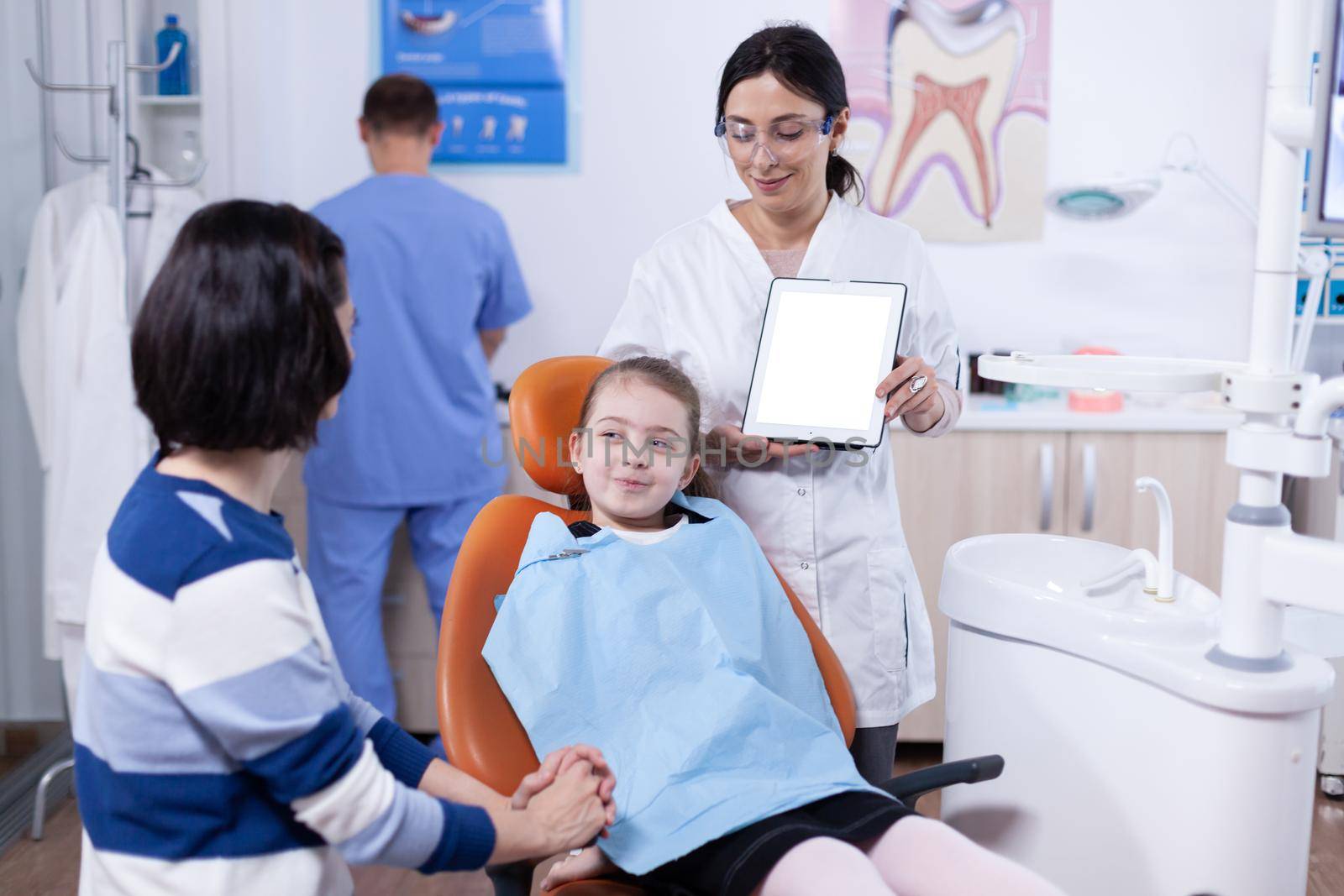 Kid wearing dental bib and doctor discussing with parent about cavity treatment. Stomatolog explaining teeth prevention to mother and child holding tablet pc with copy space available.