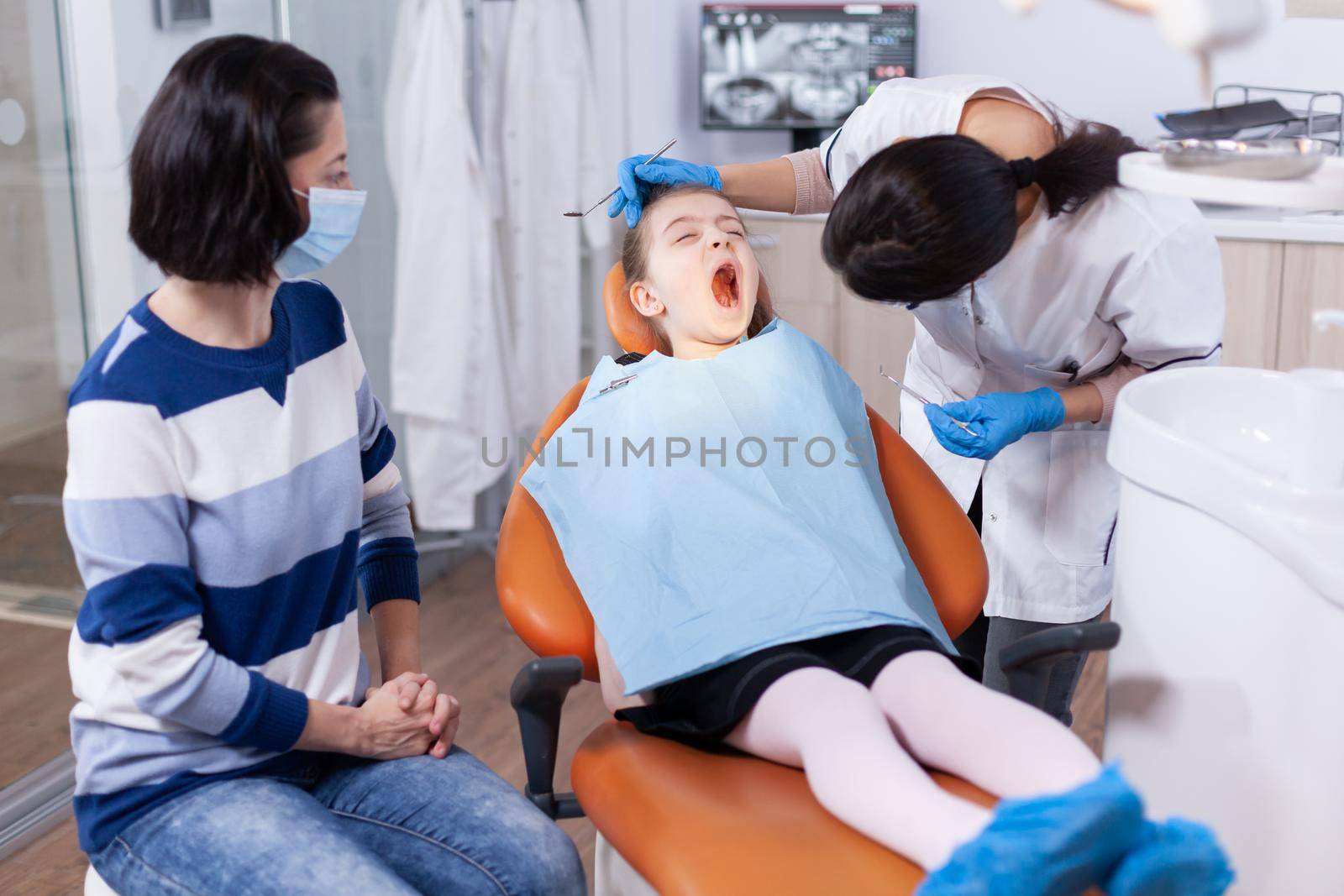 Little kid with mouth open wearing bib sittingon dental chair and dentist looking for caries Dentistry specialist during child cavity consultation in stomatology office using modern technology.