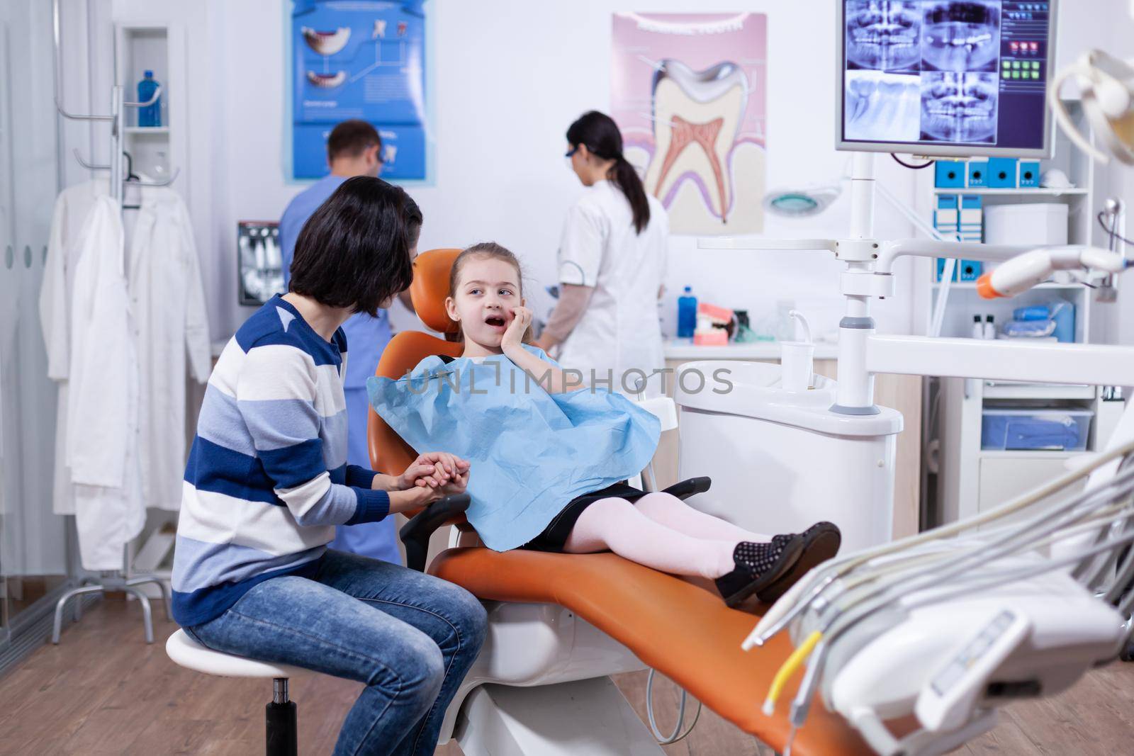 Little girl in dentist office touching mouth with hand and painfull expression. Child with her mother during teeth check up with stomatolog sitting on chair.