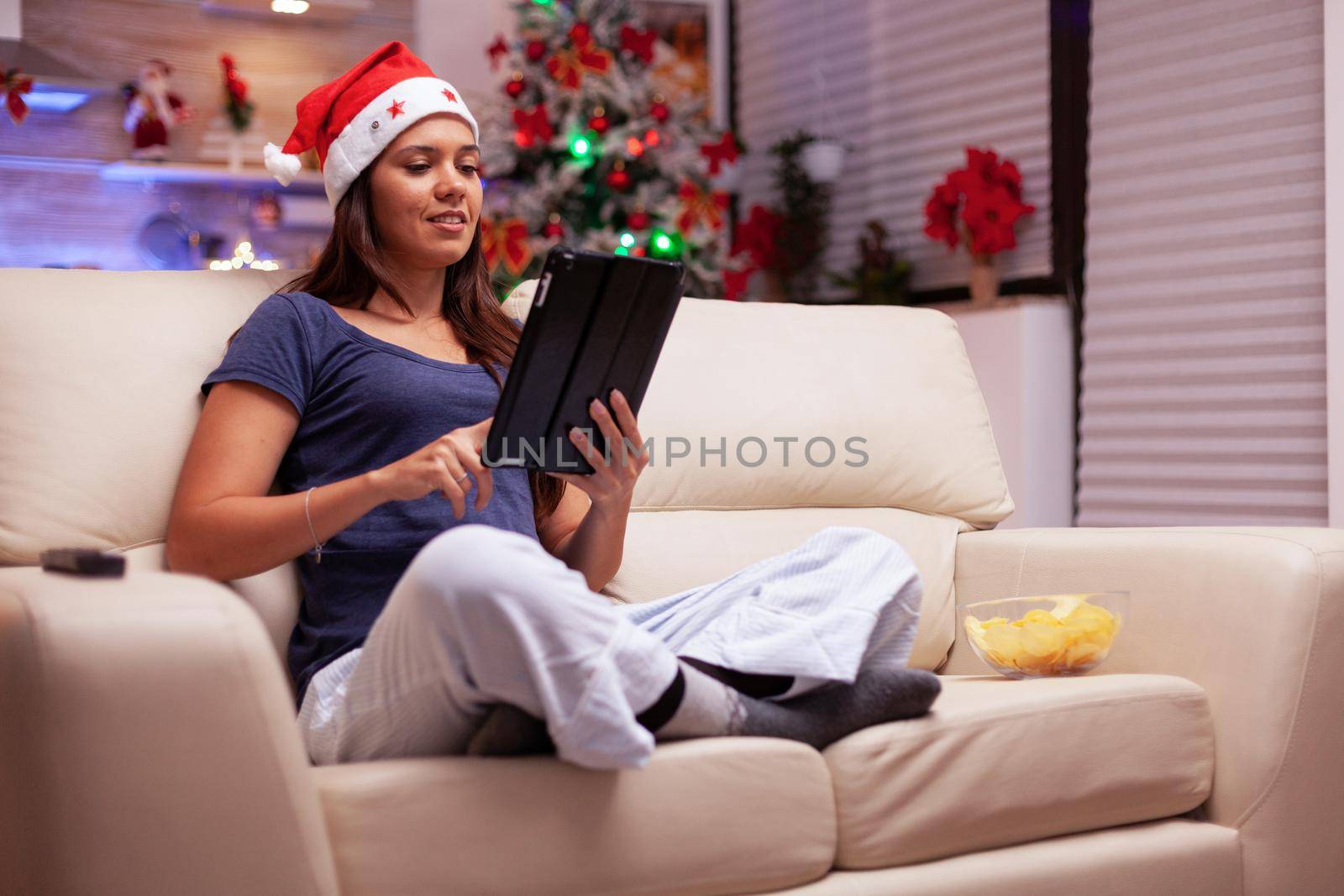 Woman sitting in lotus position on sofa browsing on social media writing xmas email messaging with friends using tablet computer. Adult person celebrating christmas season enjoying winter holiday