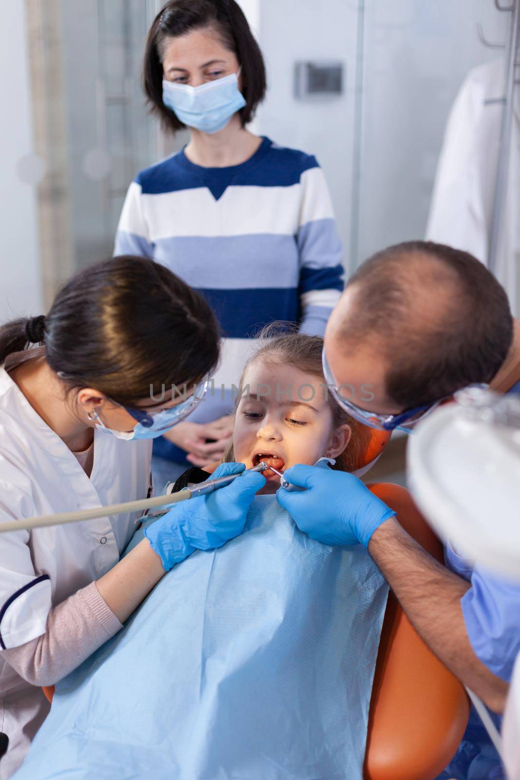 Dentist examining little girl's tooth with assistant help wearing uniform. Mother with her kid in stomatology clinic for teeth examine using modern instruments.