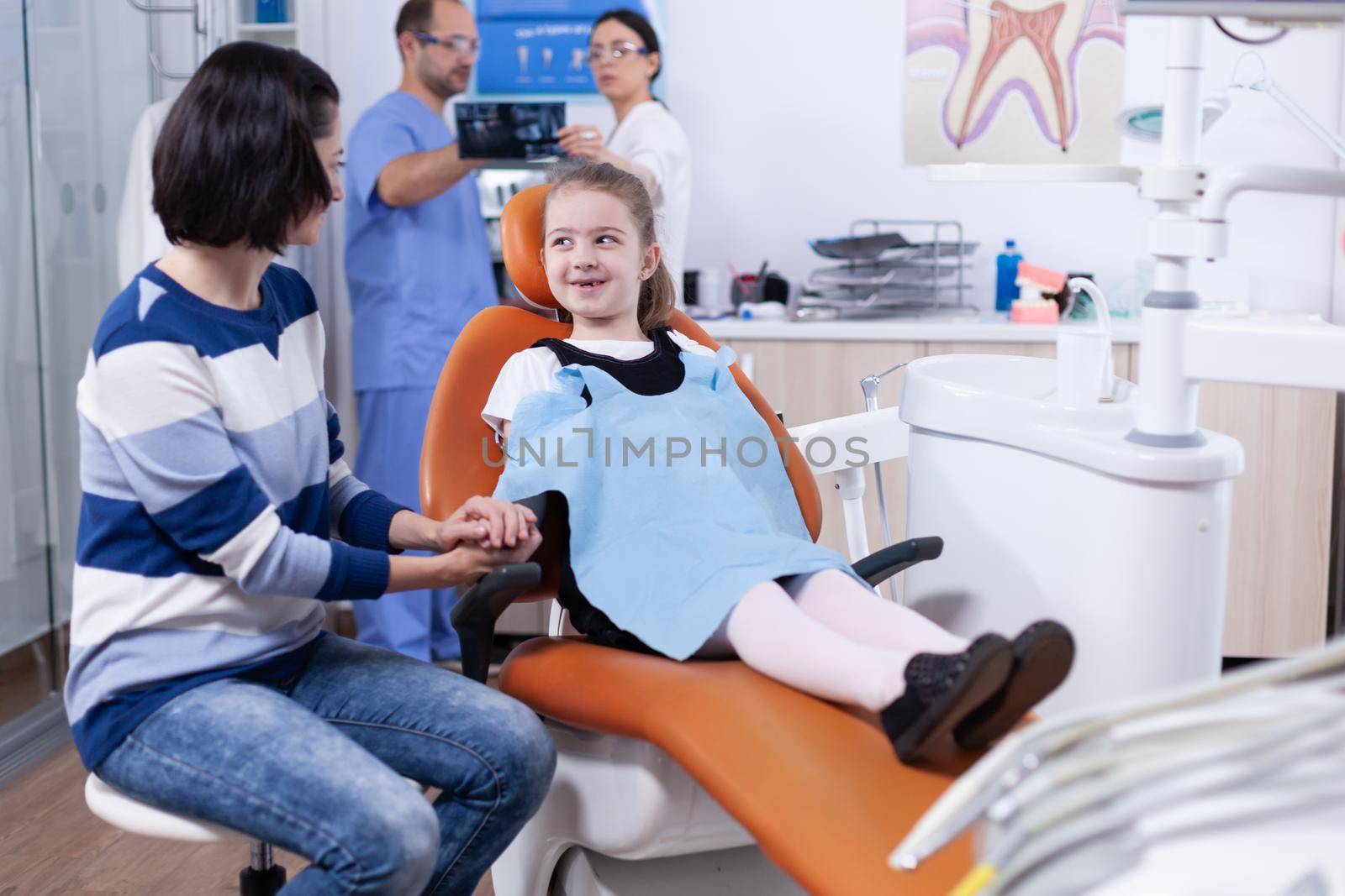 Little girl with missing tooth wearing dental bib waiting for examination in dentist office. Child with her mother during teeth check up with stomatolog sitting on chair.