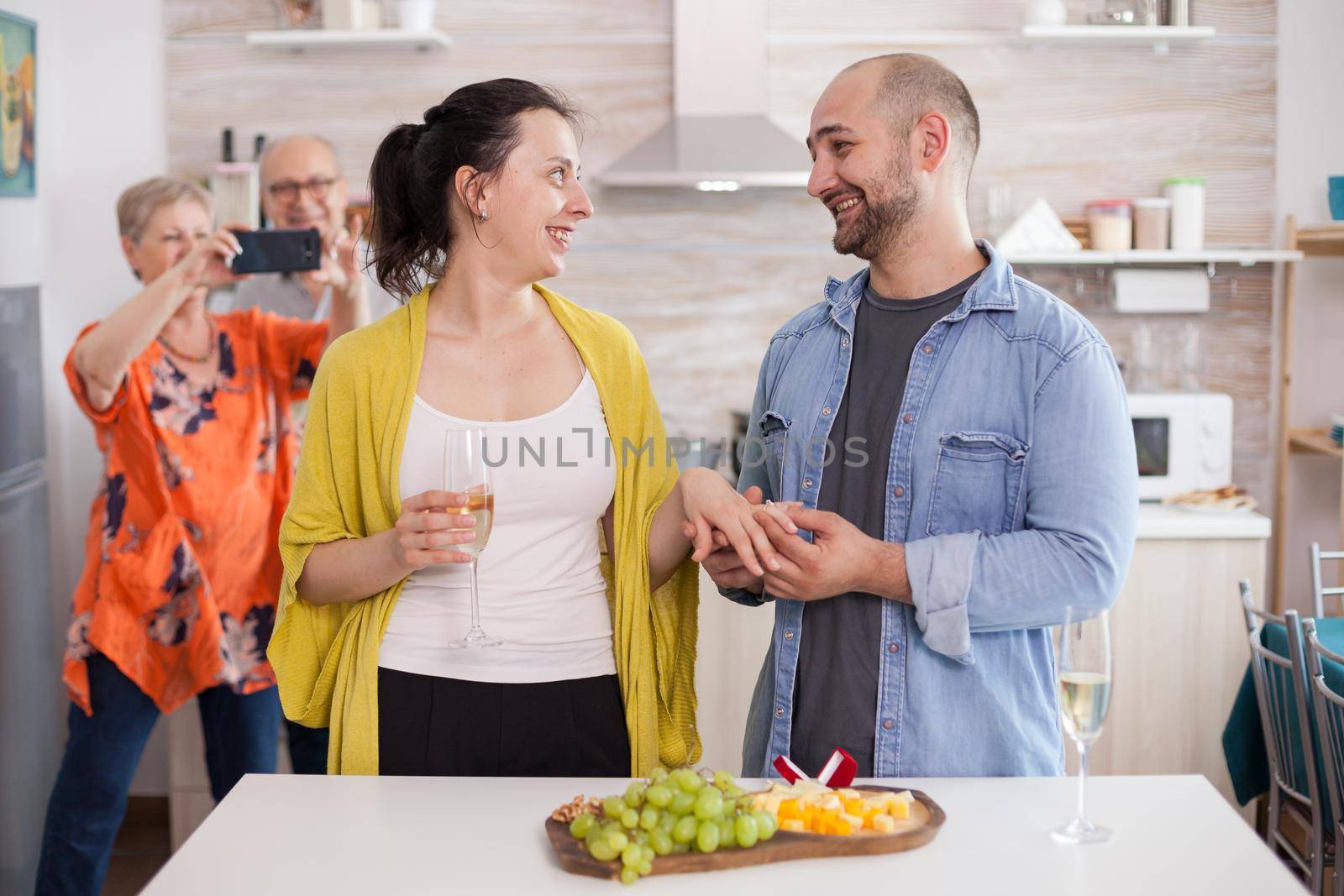 Young man smiling at wife while putting engagement ring on her finger in front of parents during family lunch.