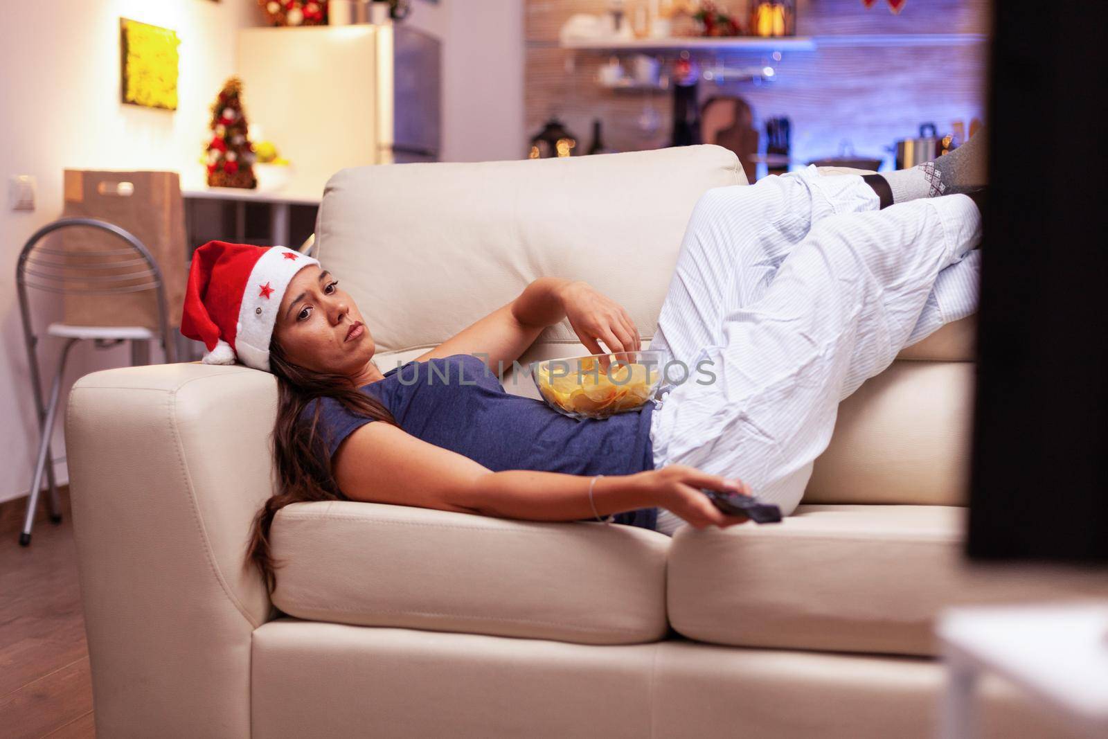 Woman relaxing on couch eating snacks while watching xmas comedy movie on tv during christmastime in x-mas decorated kitchen. Adult person enjoying winter season celebrating christmas holiday