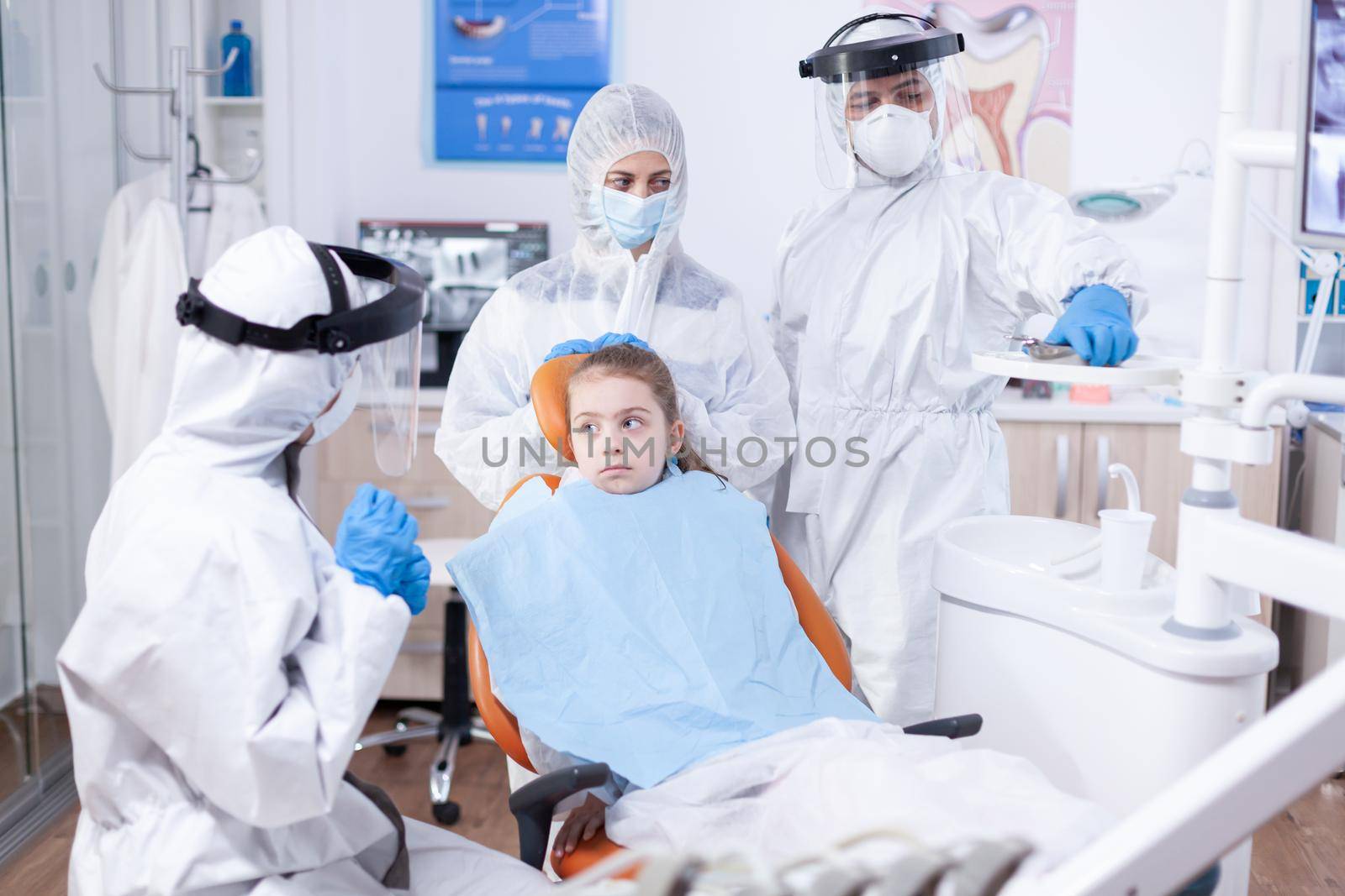Dentist explaining dental problem dressed in protection suit in the course of coronavirus pandemic. Stomatologist during covid19 wearing ppe suit doing teeth procedure of child sitting on chair