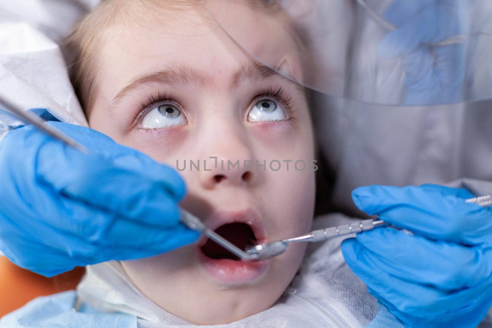 Close up of kid getting caries treatment from dentist in the course of covid19 outbreak. Dentist in coronavirus suit using curved mirror during teeth examination of child.