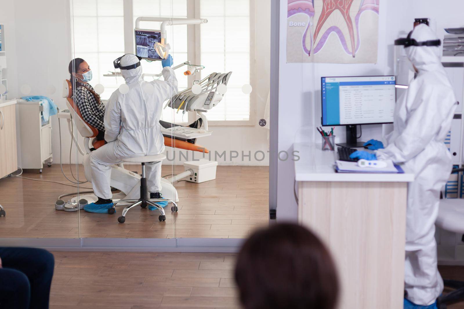 Stomatologist in protective suit asking for patient dental x-ray examining teeth problems during coronavirus pandemic in modern clinic. Medical team wearing coverall, face shield, mask and gloves.