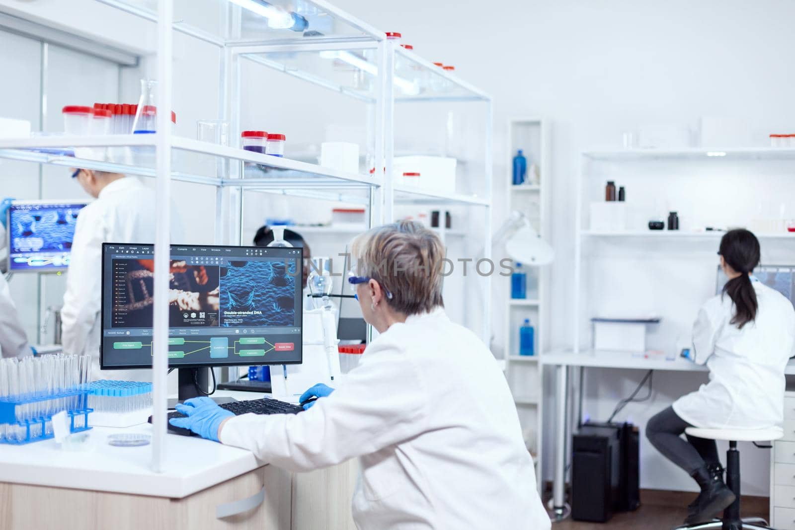 Elderly researcher working to find a cure during global pandemic. Senior scientist in pharmaceuticals laboratory doing genetic research wearing lab coat with team in the background.