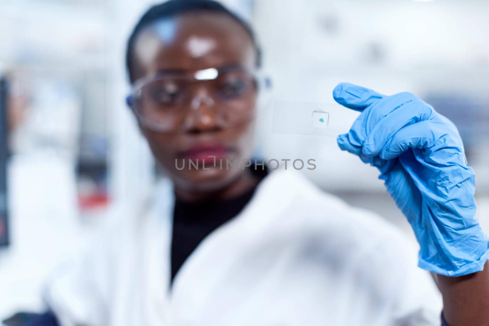 Sample glass slide in focus while chemist is looking at it during lab investigation. African healthcare scientist in biochemistry laboratory wearing sterile equipment.