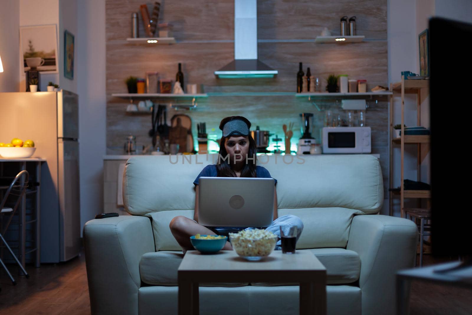 Woman with eye mask using laptop at night while watching tv and eating snacks. Happy person in pijamas sitting on sofa reading writing searching browsing on notebook using internet checking mails
