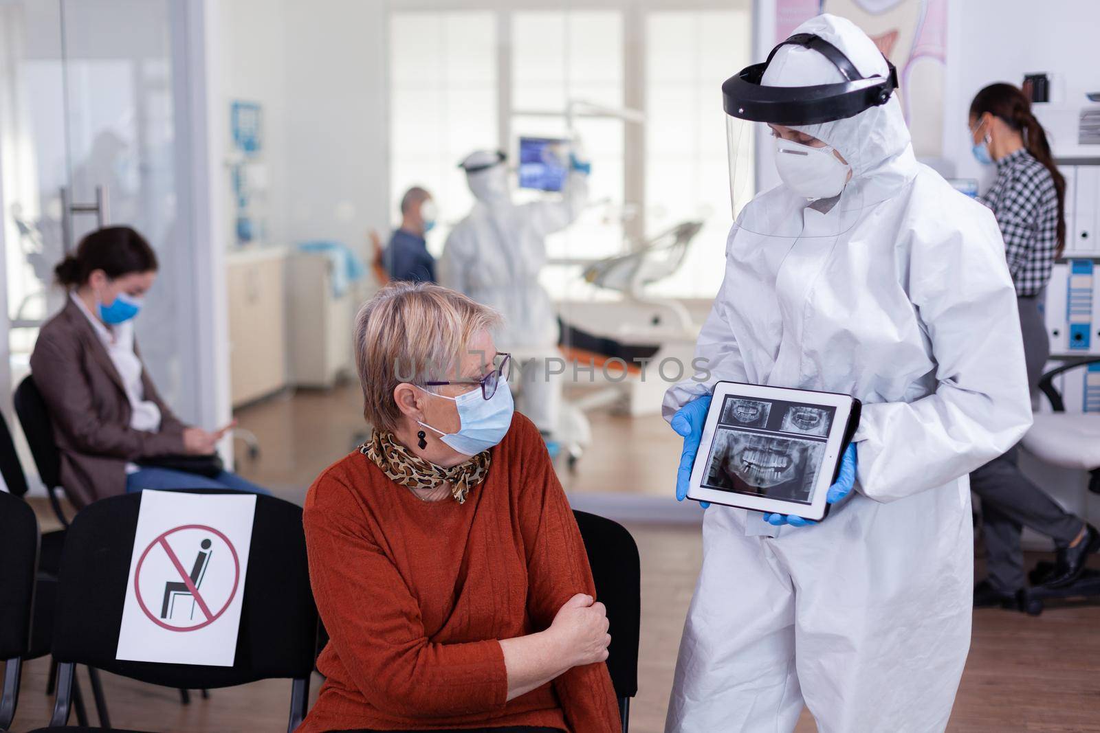 Dentist doctor with face shield pointing on tablet display explaining dental x-ray to senior patient during global pandemic. Nurse wearing protection suit, overall, mask and gloves, new normal.