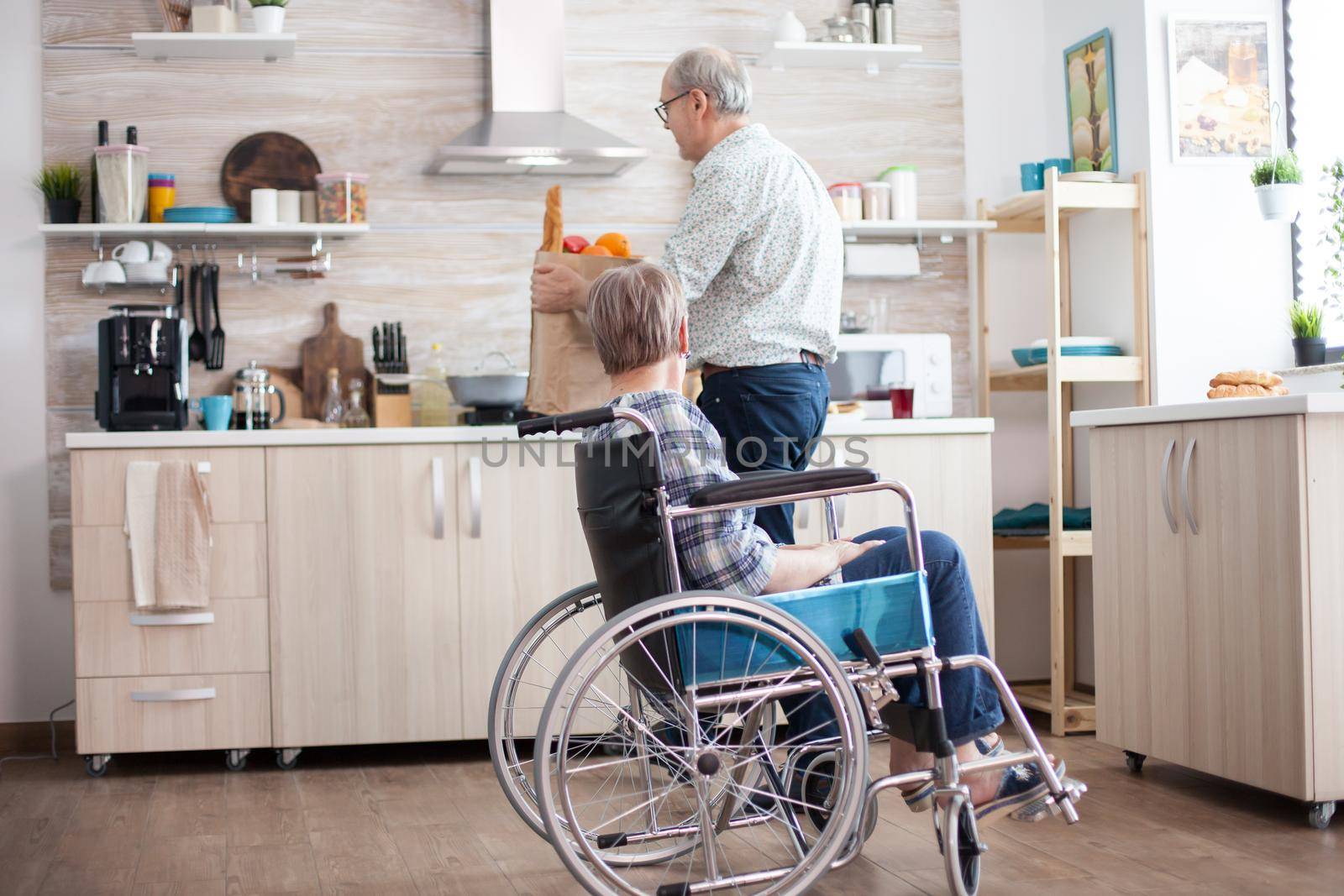 Husband helping disabled senior woman in wheelchair with grocery paper bag. . Mature people with fresh vegetables from market. Living with disabled person with walking disabilities