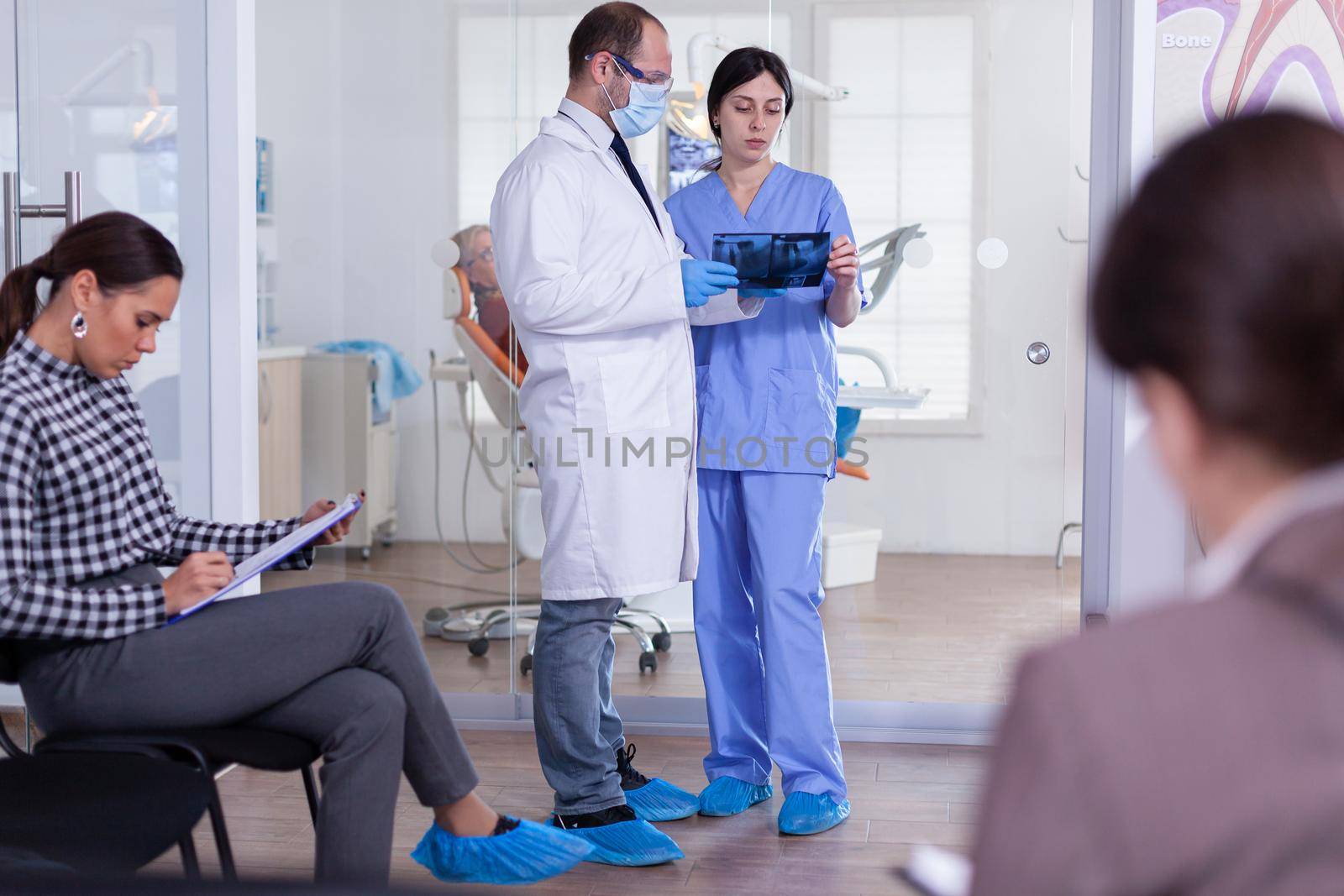 Dentist in stomatology waiting area holding patient x-ray explaining diagnosis to nurse in a crowded hallway with patient filling form. Dentistiry staff in reception.