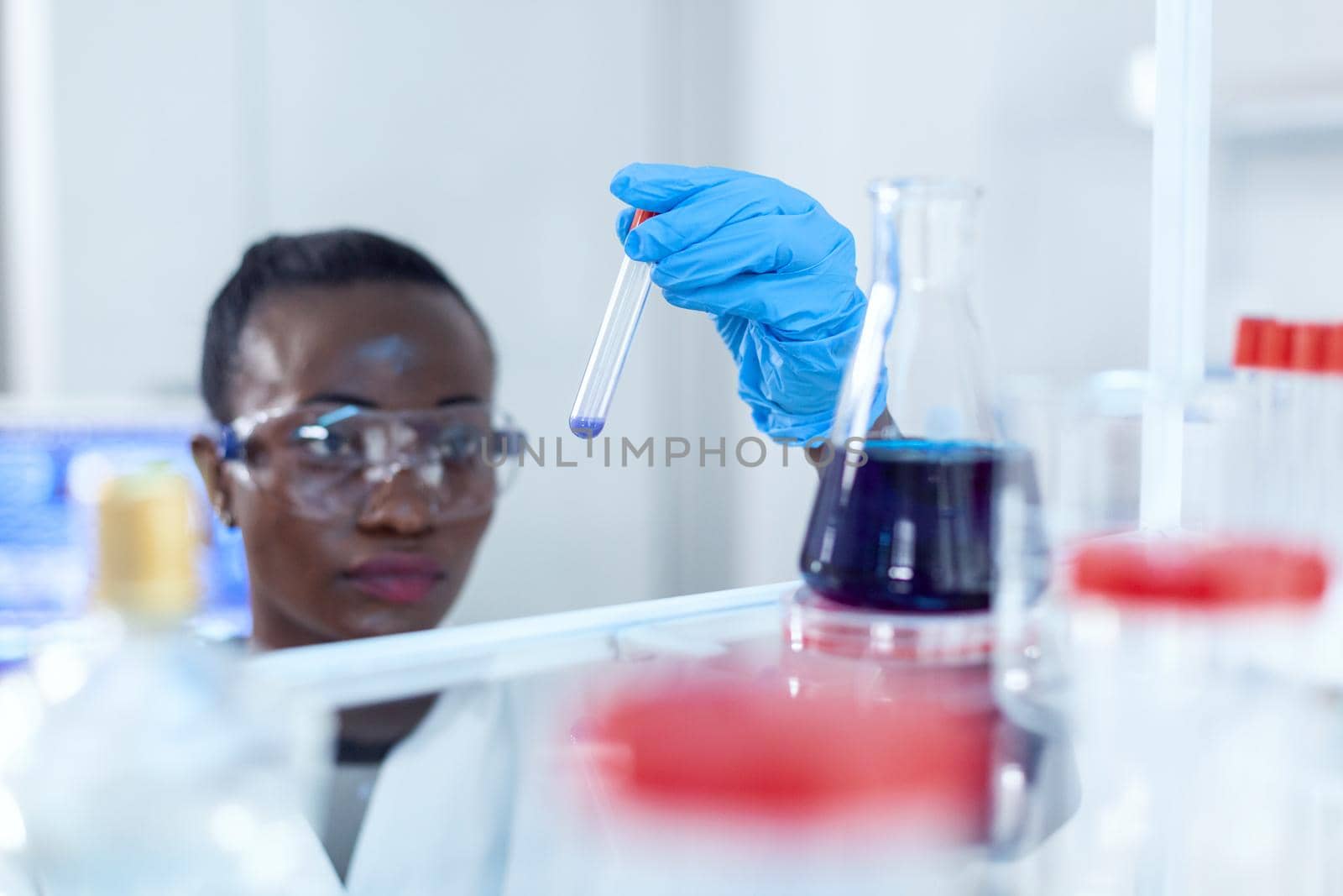 African scientist staring at sample blue liquid in test tube chemical. Black researcher in sterile laboratory conducting pharmacology experiment wearing coat.