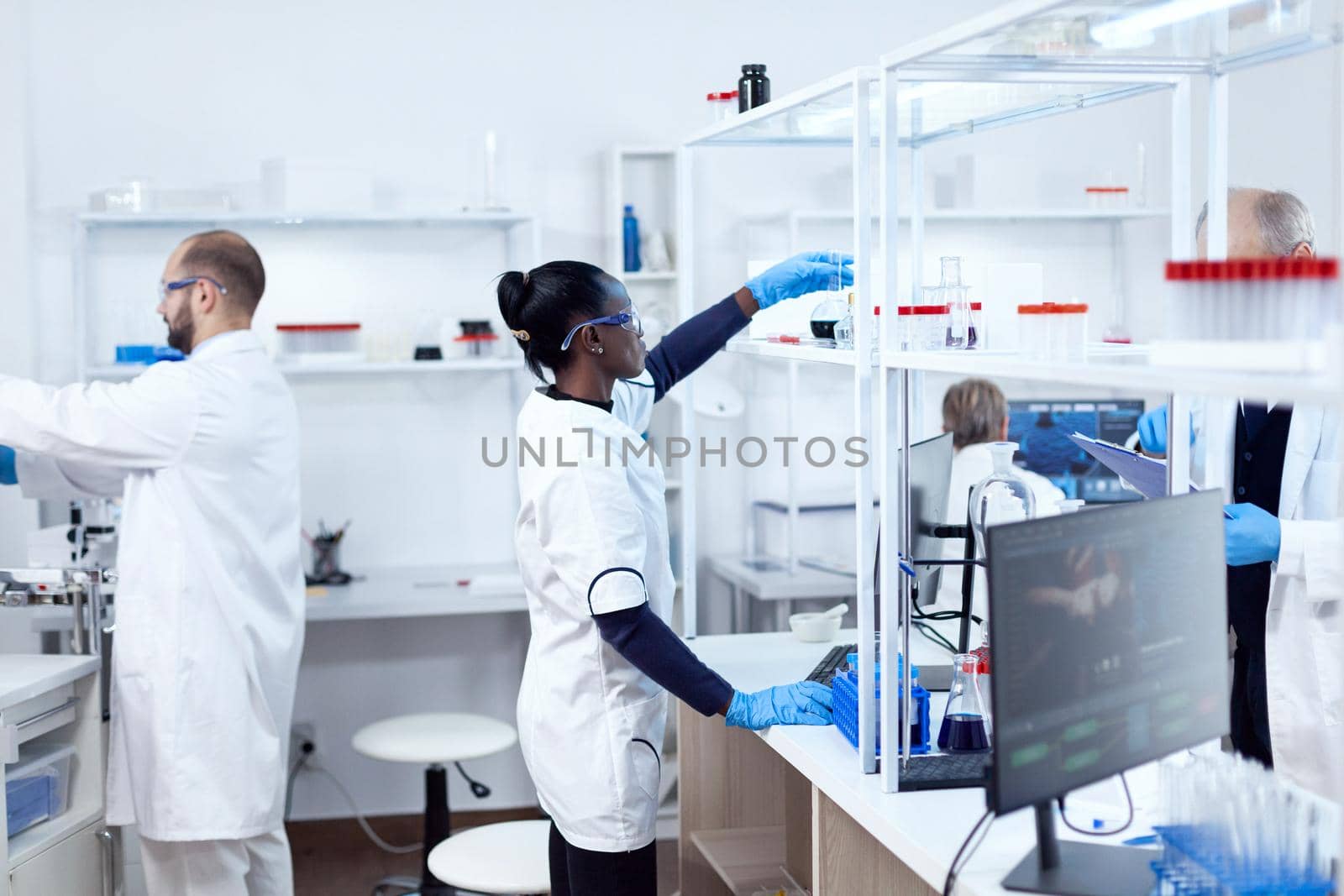 African in biotechnology laboratory reaching for glass flask from shelf. Multiethnic team of researchers working in microbiology lab testing solution for medical purpose.