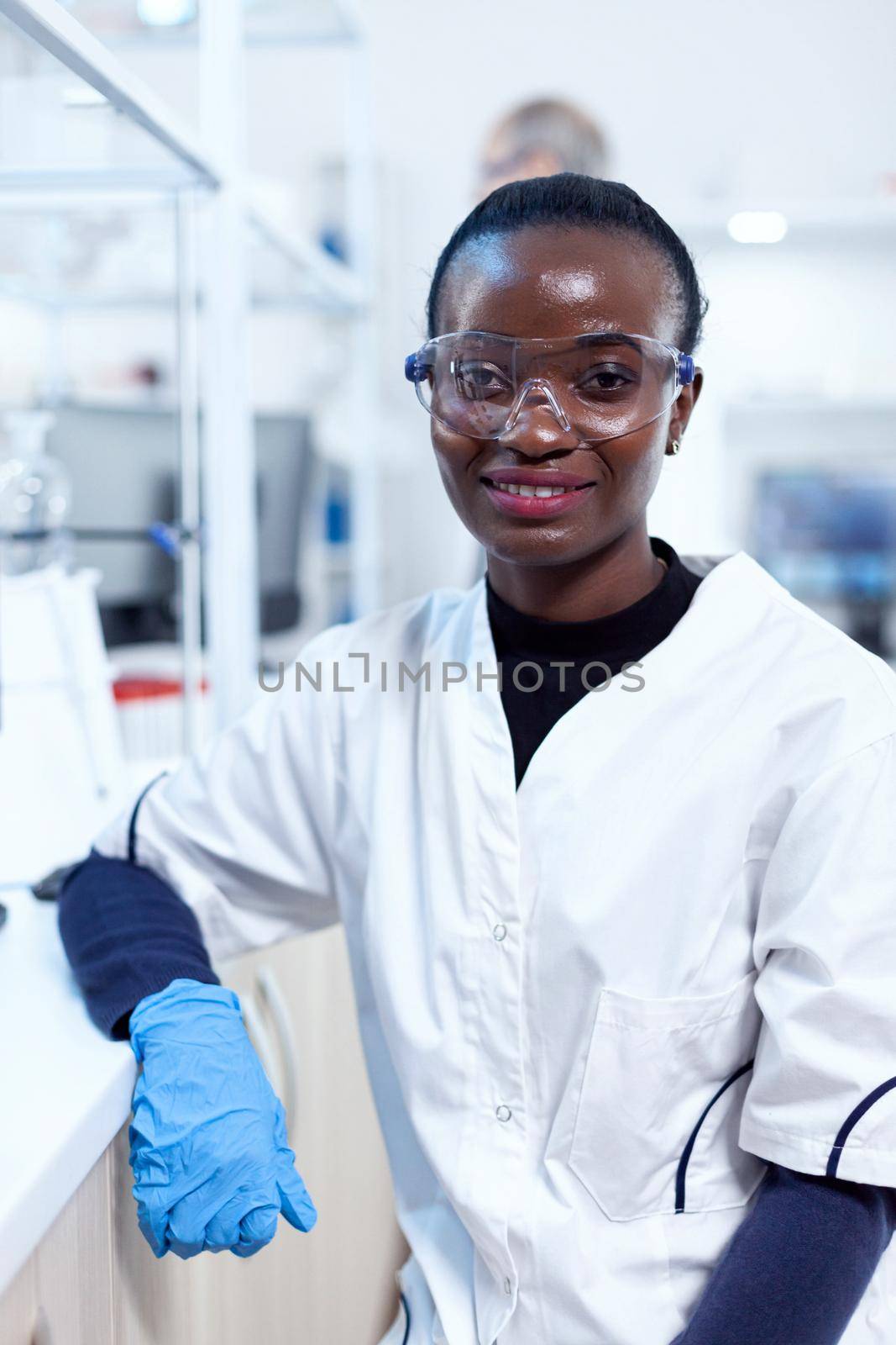African scientist wearing protective glasses in medical laboratory looking at camera. Multiethnic team of researchers working in microbiology lab testing solution for medical purpose.