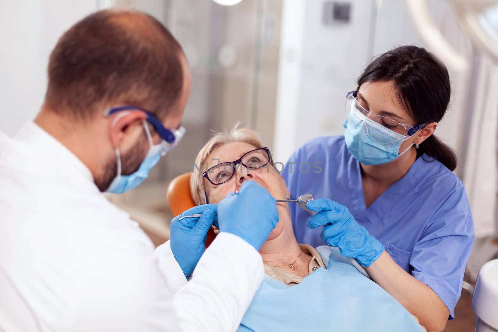 Stomatolog with assistant putting dental seal on senior woman teeth. Elderly patient during medical examination with dentist in dental office with orange equipment.