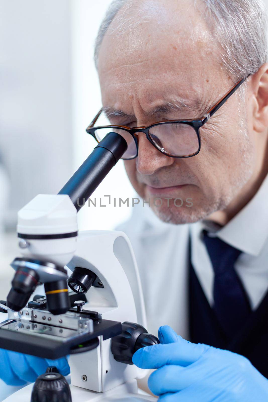Elderly aged scientist in modern facility looking through a microscope during health care study. Chemist researcher in sterile lab doing experiments for medical industry using modern technology.