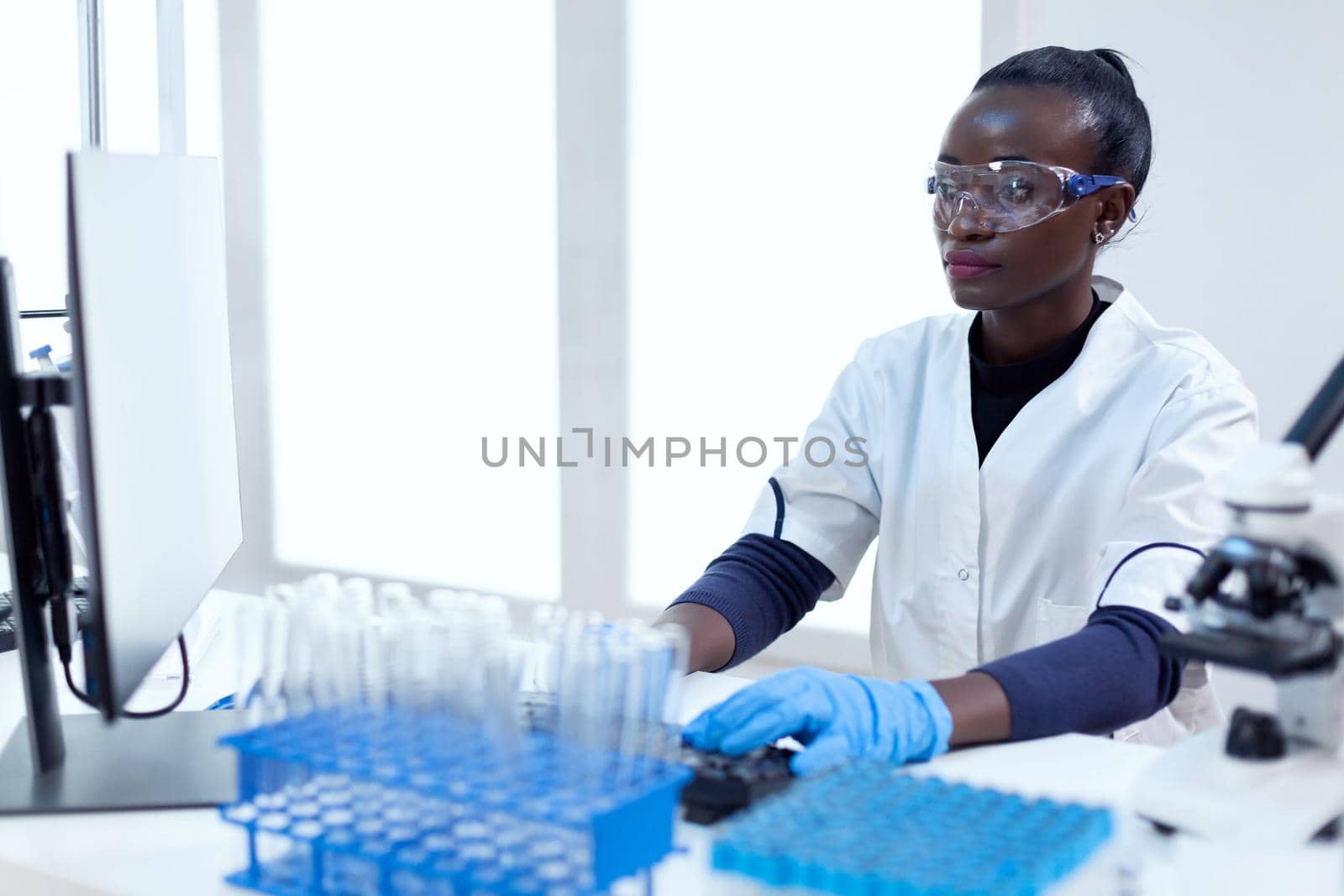 African biotechnology researcher works in bright modern laboratory using computer. Multi ethnic healthcare scientist in biochemistry facility wearing sterile equipment.