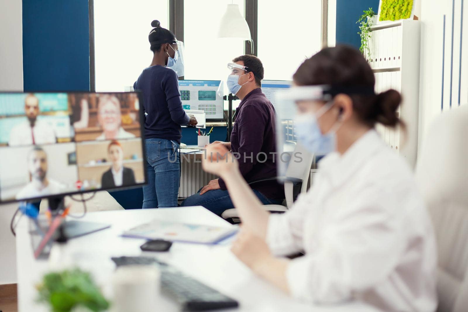 Man discussing with african coworker about a project at workplace wearing face mask against covid. Entrepreneur having videocall while colleagues working respecting social distance during global pandemic.