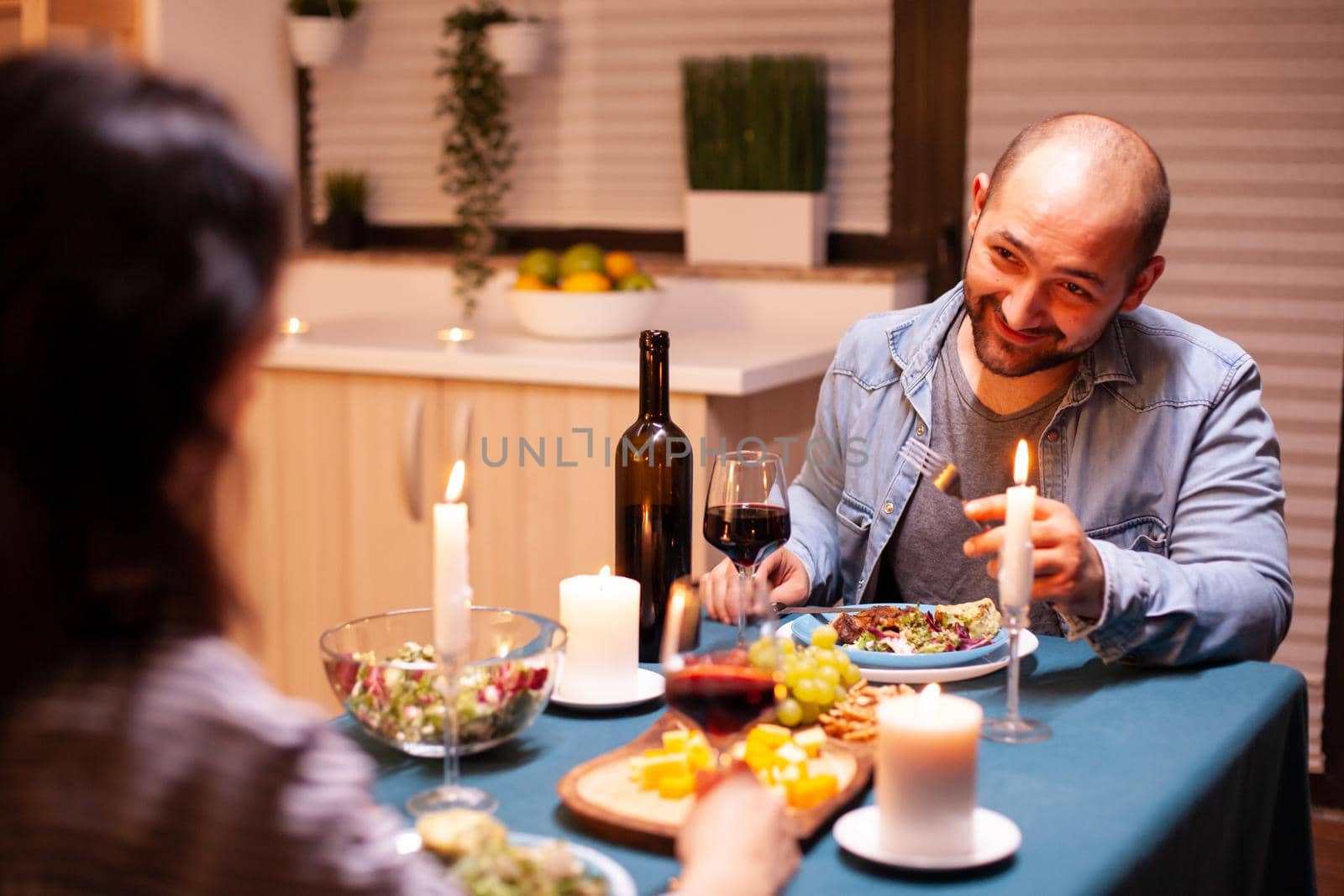 Boyfriend looking with love at wife and eating tasty food during dinner in kitchen. Talking happy sitting at table dining room, enjoying the meal at home having romantic time at candle lights.