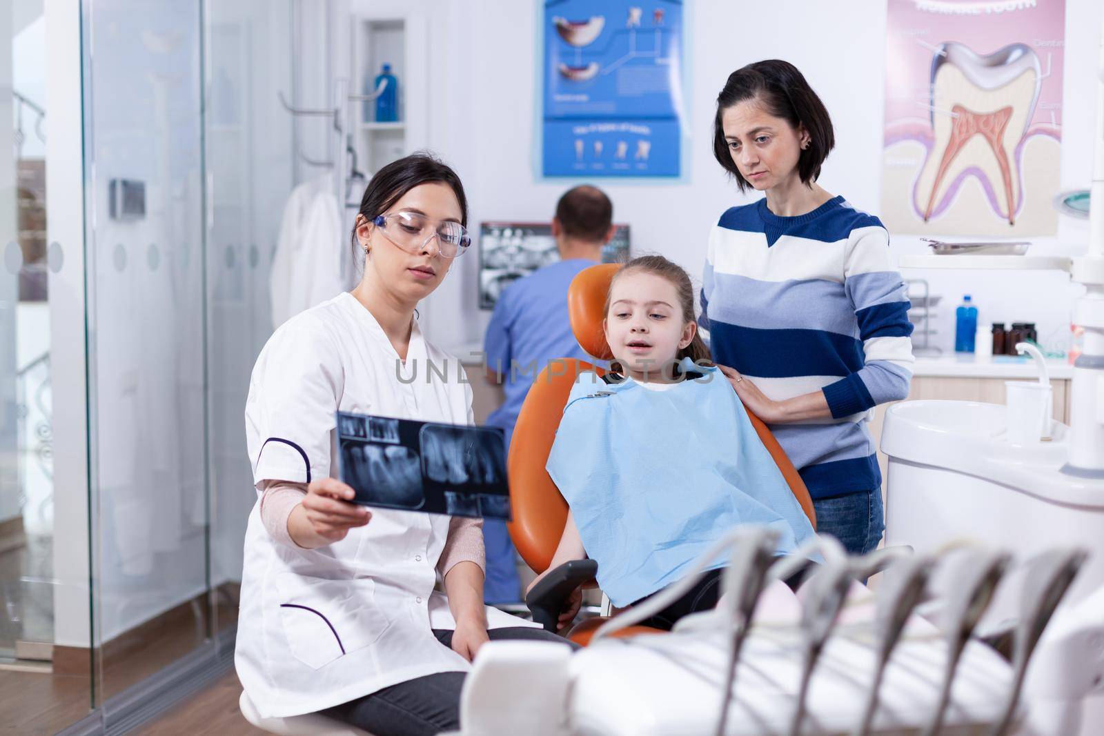 Dentist examining little kid radiography sitting on chair wearing dental bib. Stomatologist explaining teeth diagnosis to mother of child in health clinic holding x-ray.