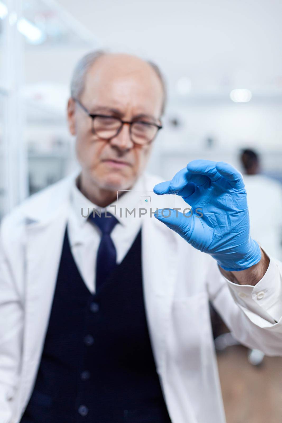 Concentrated senior scientist looking at glass slide with sample wearing protection gloves. Elderly researcher in sterile lab looking on microscope slide wearing lab coat.