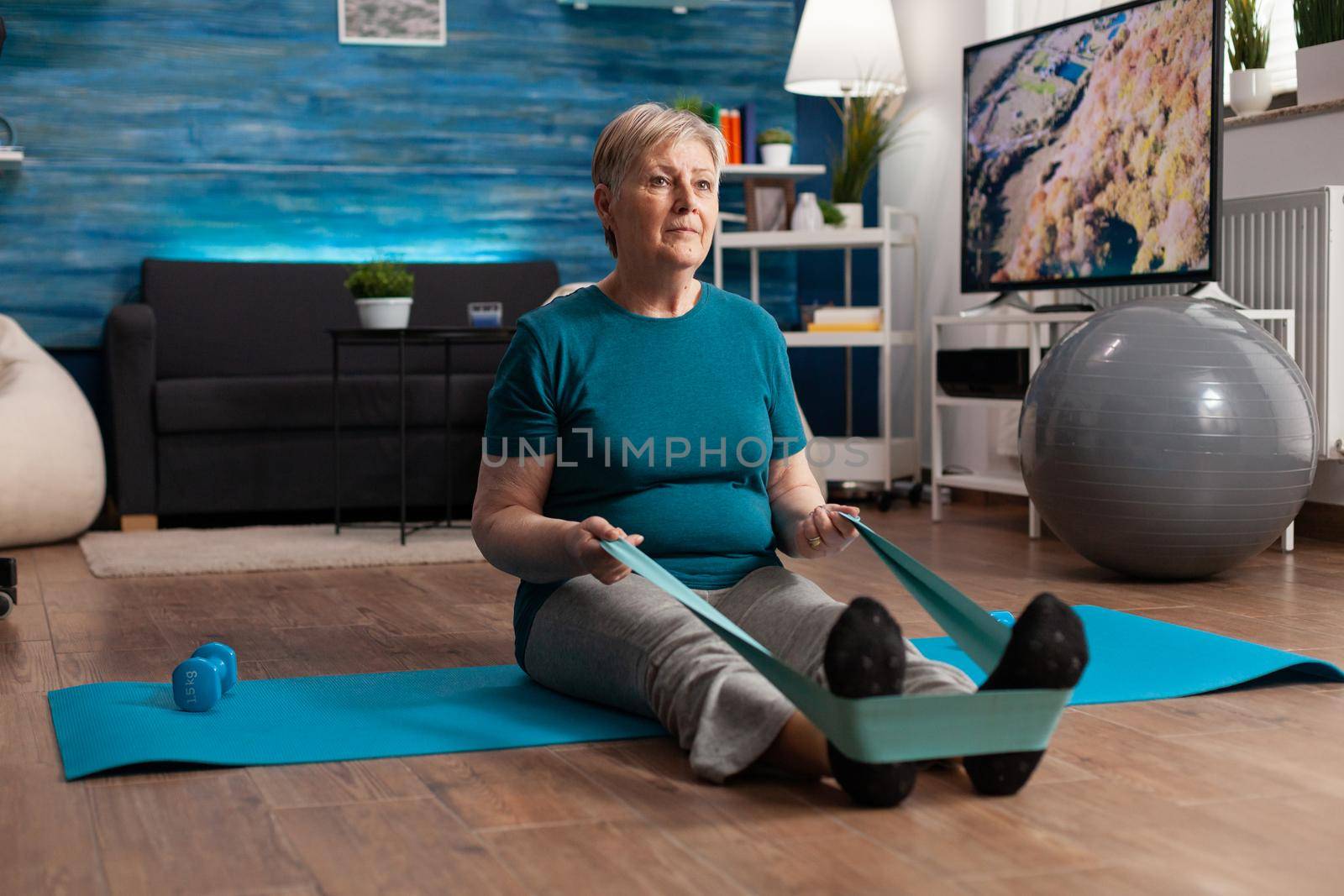 Retirement senior woman sitting on yoga mat stretching legs muscles using stretch elastic band training body resistance. Pensioner in sportswear slimming weight during muscle training in living room