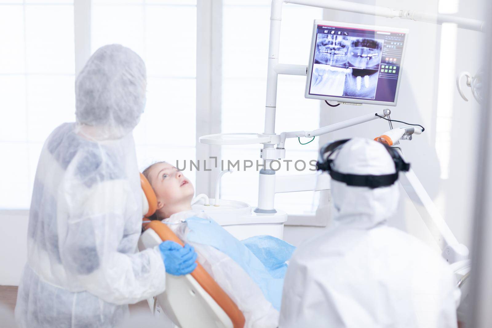 Little girl in ppe looking at her mother in the course of dental examination. Stomatolog in protectie suit for coroanvirus as safety precaution looking at digital child teeth x-ray during consultation.