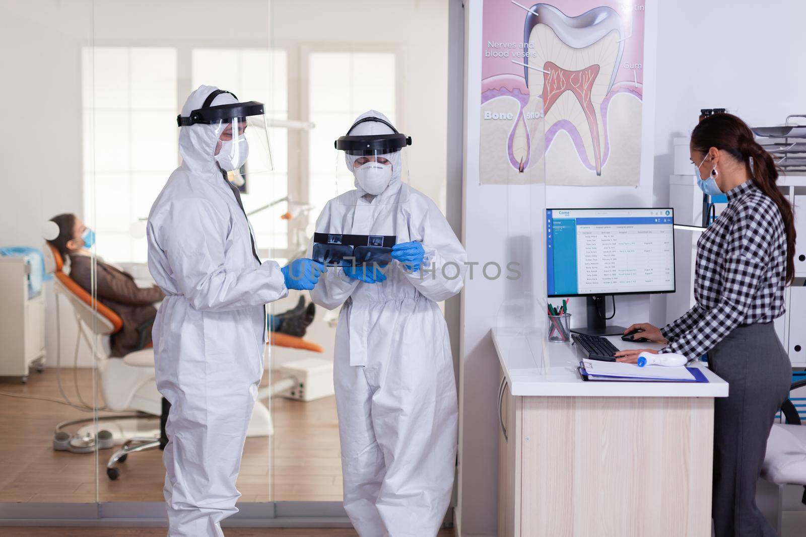 Stomatology team dressed up in ppe suit during global pandemic with coronavirus in dental reception holding patient x-ray, keeping social distancing. Receptionist with face mask as prevention.